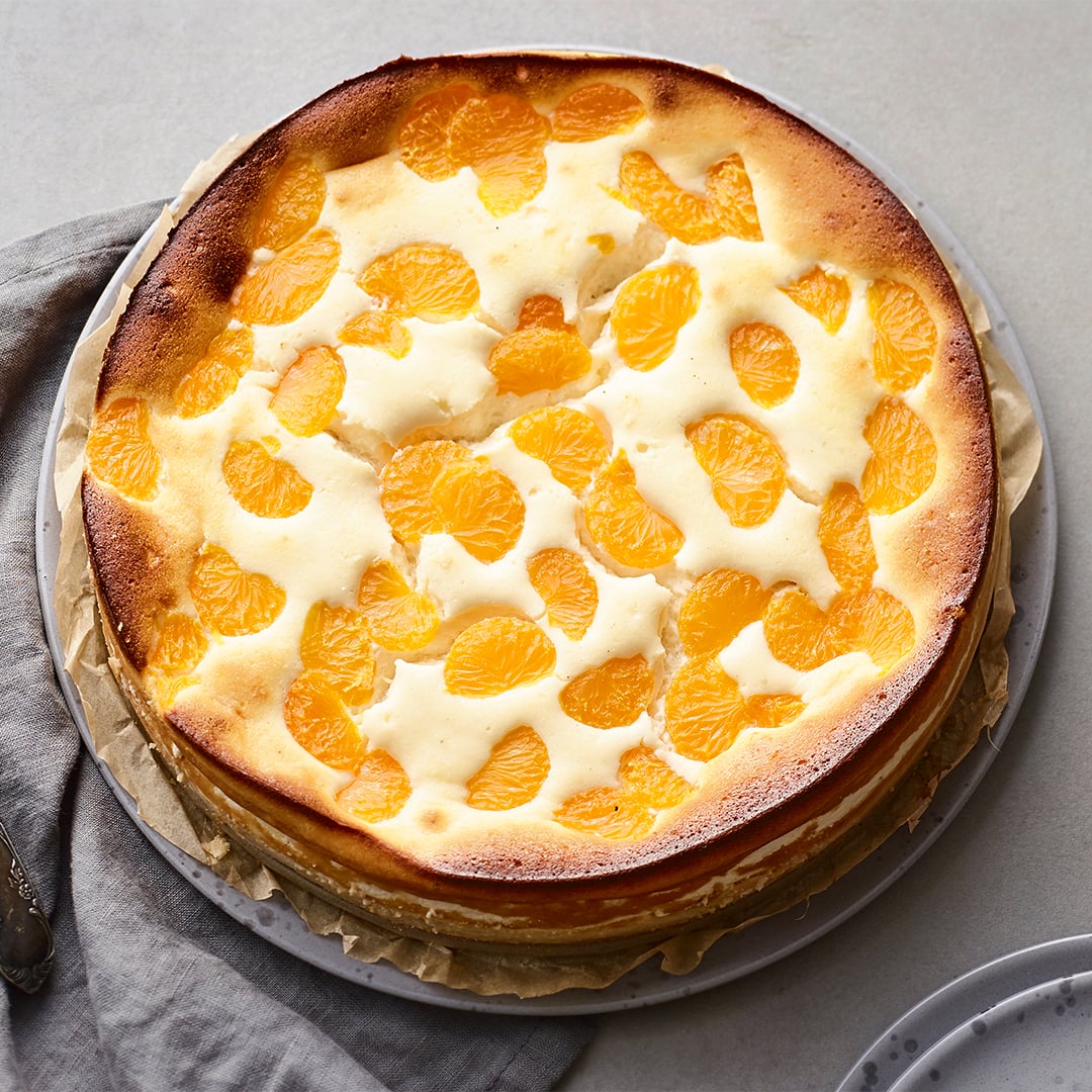 Luftiger Käsekuchen mit gelbgold gebackenem Rand, getoppt mit Mandarinenstückchen, auf einem steingrauen Teller