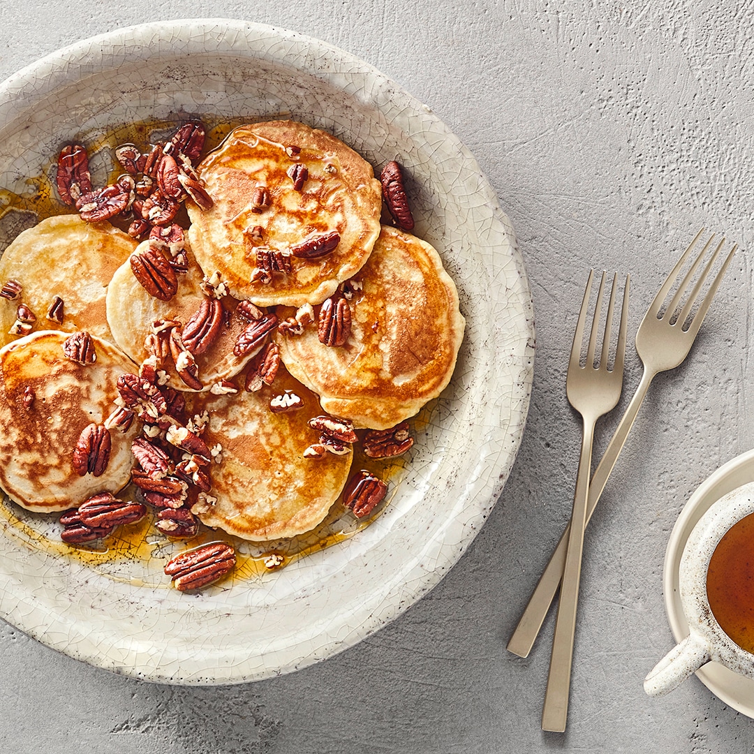 Ein Teller mit kleinen runden Pfannkuchen, braunen Pekannüssen und goldener Flüssigkeit