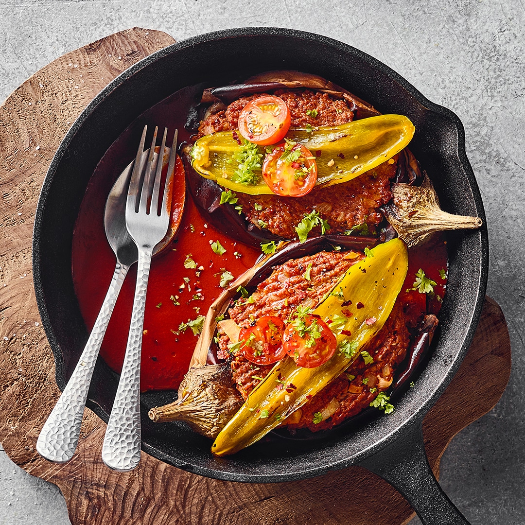 Eine Pfanne mit zwei gefüllten Auberginenhälften in roter Sauce, garniert mit grünen Kräutern, gelben Paprikaschoten und Tomaten