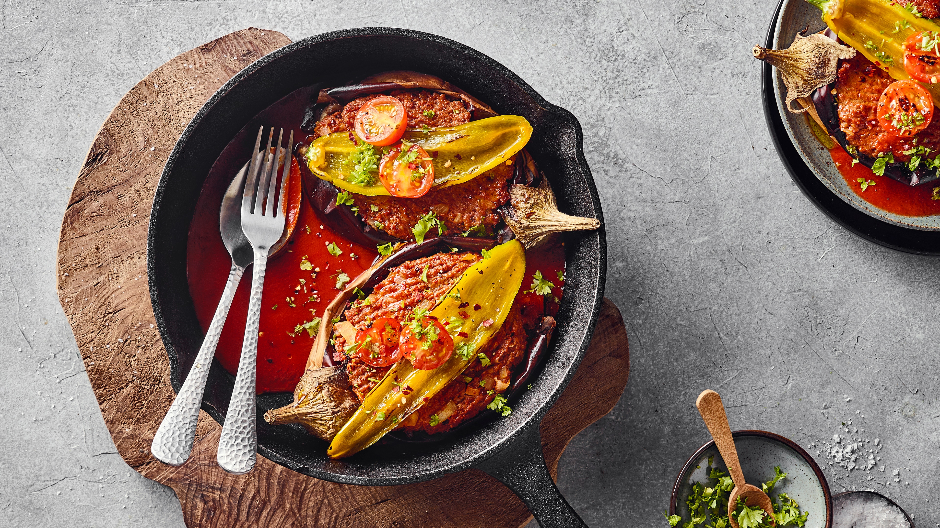 Eine Pfanne mit zwei gefüllten Auberginenhälften in roter Sauce, garniert mit grünen Kräutern, gelben Paprikaschoten und Tomaten