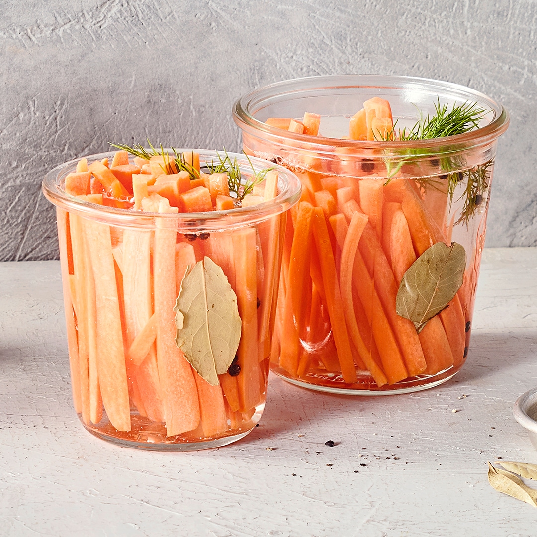 Zwei Gläser mit orangefarbenen Karottenstreifen in Flüssigkeit, darin grüne Kräuter und ganze Lorbeerblätter