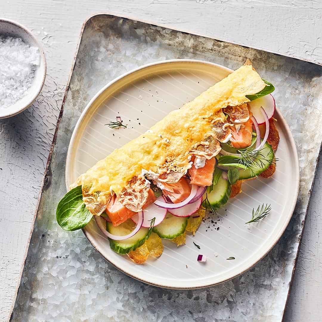 Ein Klappsandwich mit einer knusprigen Kartoffelkäsekruste, Gemüse und Stremellachsstücken auf einem runden Teller.