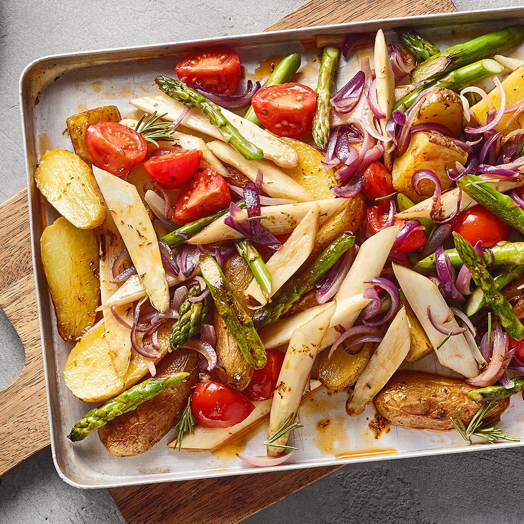 Ein Blech mit gebratenen Kartoffelstücken, grünen Spargelstangen, weißen Wurzelstreifen, roten Tomatenhälften und violetten Zwiebelringen.