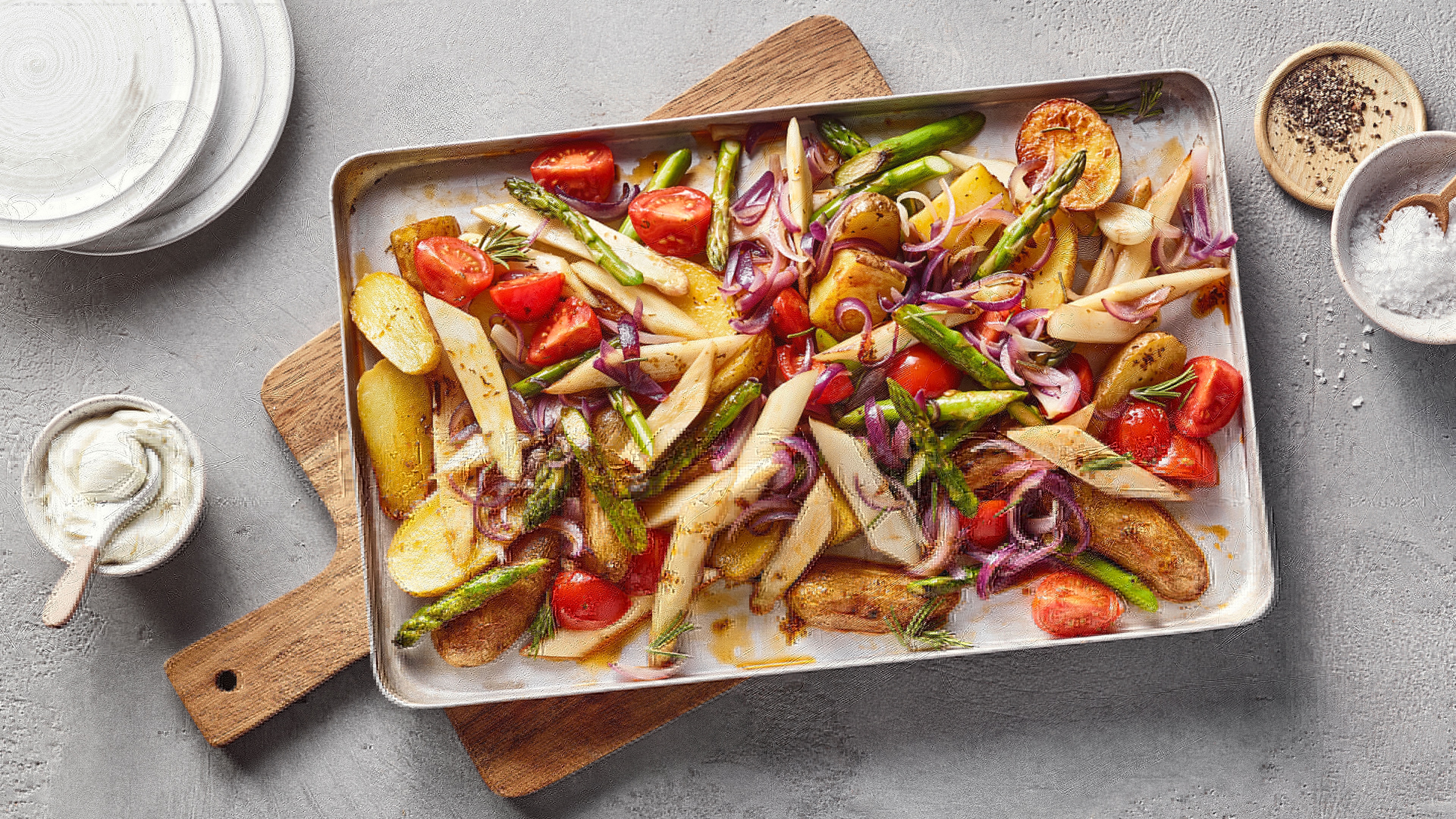 Ein Blech mit gebratenen Kartoffelstücken, grünen Spargelstangen, weißen Wurzelstreifen, roten Tomatenhälften und violetten Zwiebelringen.