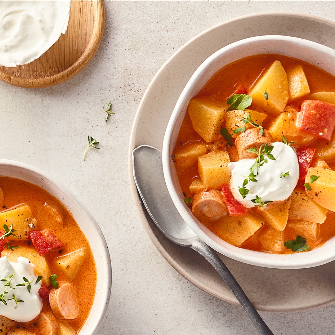 Schalen, befüllt mit einer hellen Suppe aus Kartoffeln, Paprika und Würstchen, getoppt mit weißer Creme und frischen Kräutern