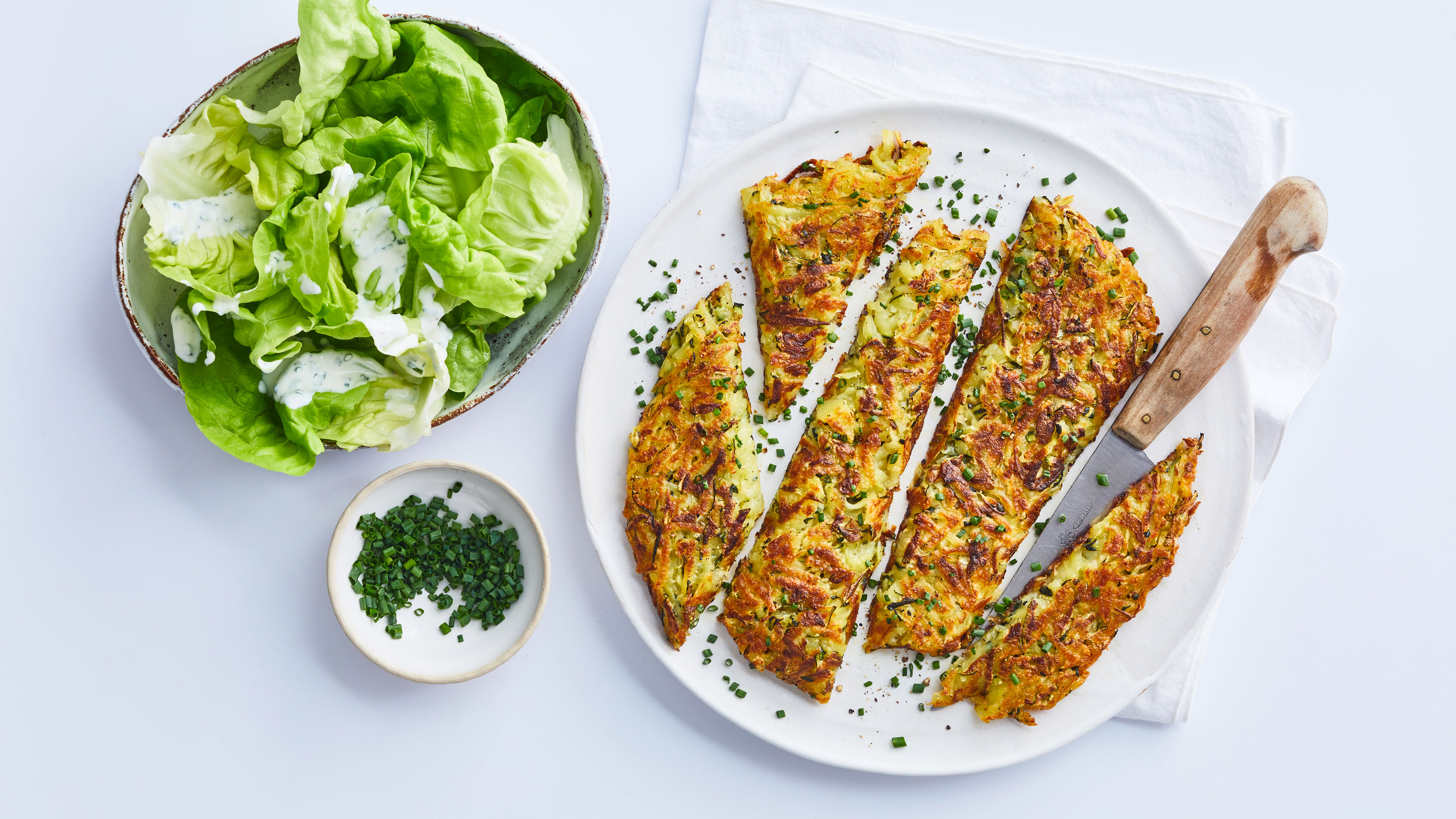 Goldbraun gebratener Zucchini-Puffer in Stücke geschnitten auf einem weißen Teller, dazu grüner Blattsalat mit Dressing und frischer Schnittlauch.