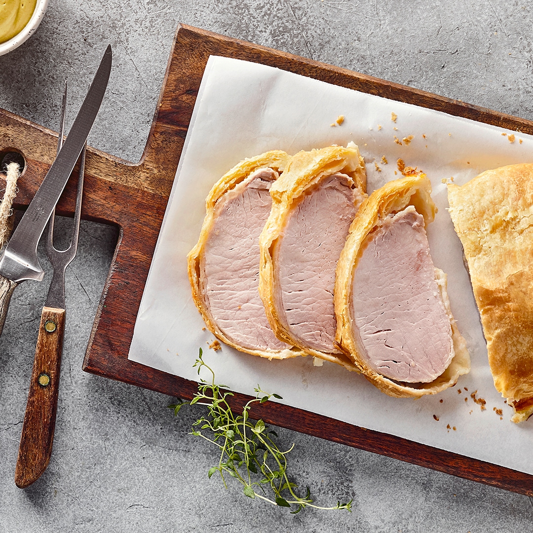 Ein großes Stück zartrosa Fleisch, ummantelt mit gelbgold gebackenem Blätterteig, auf einem rustikalen Holzbrett