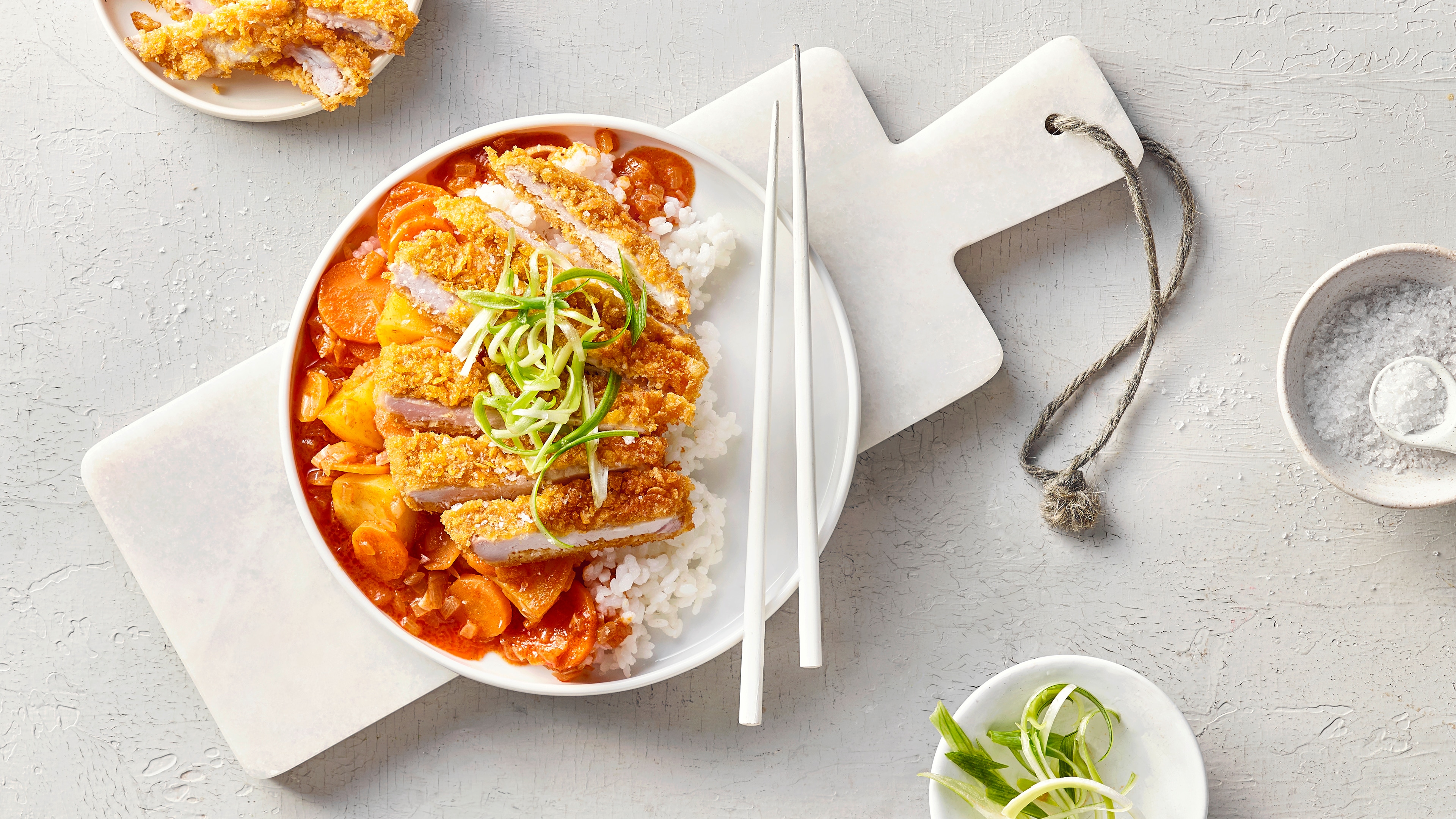 Ein Teller mit weißem Reis, darauf knusprig paniertes Fleisch in Streifen, serviert mit Gemüse in brauner Sauce und grünen Lauchstreifen