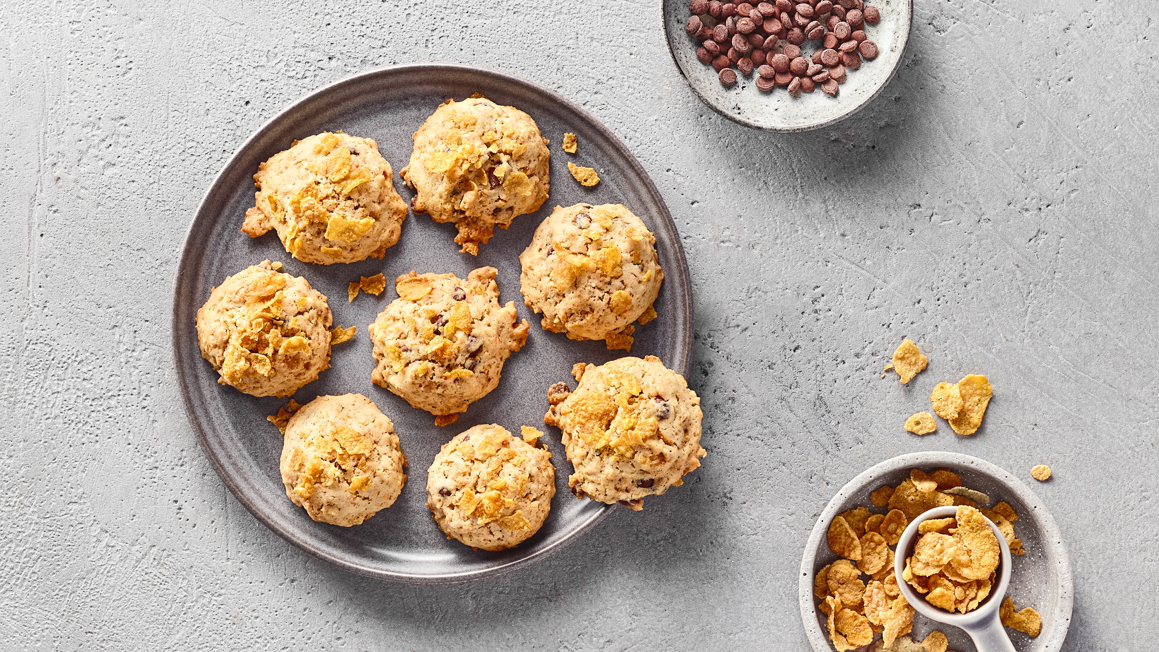 Knusprige Cookies mit Cornflakes und Schokostückchen auf einem grauen Teller, daneben Schokolinsen und Cornflakes.