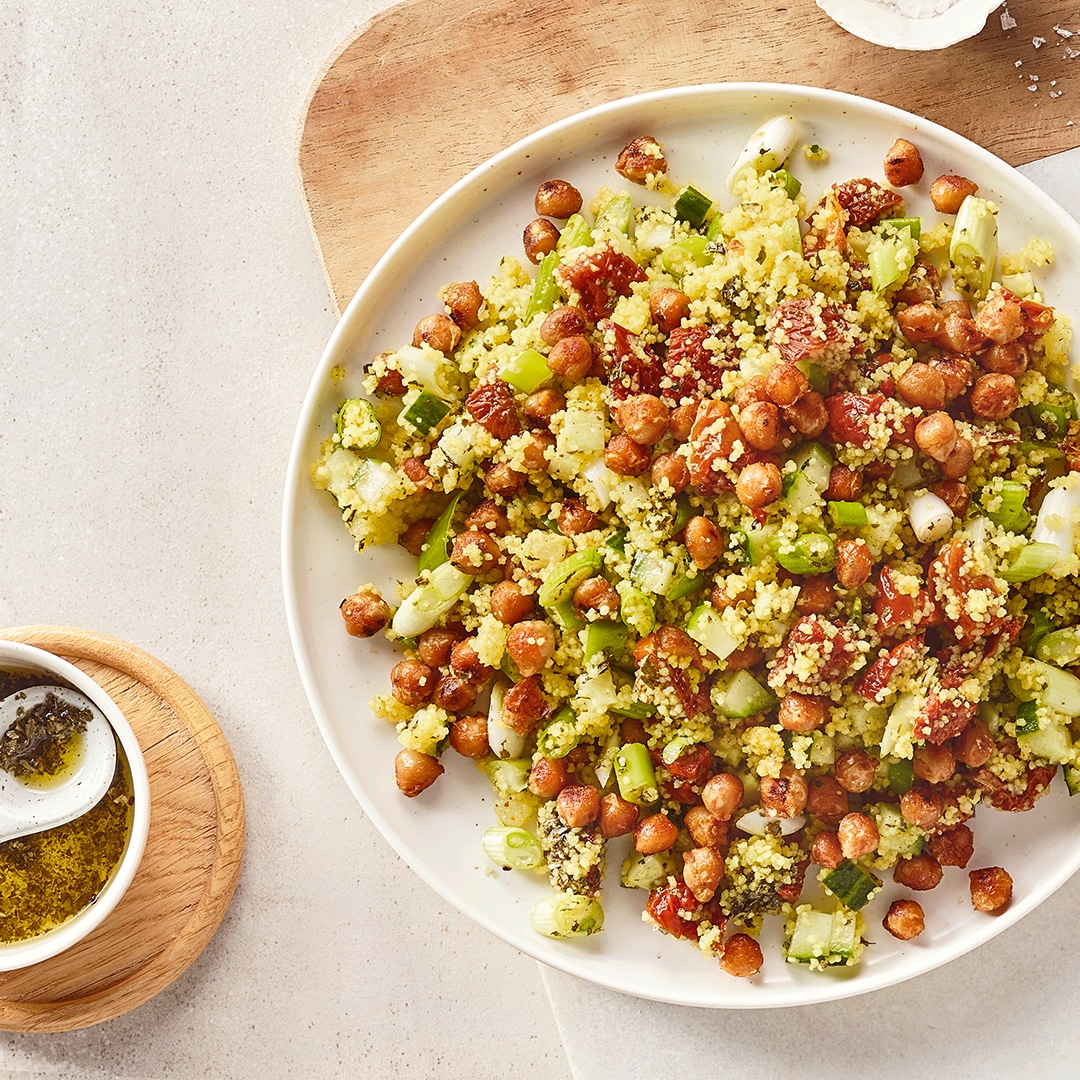 Ein Teller mit Couscous, grünen Gurkenstücken, roten Tomatenstücken und braunen Kichererbsen, daneben eine Schale mit Öl
