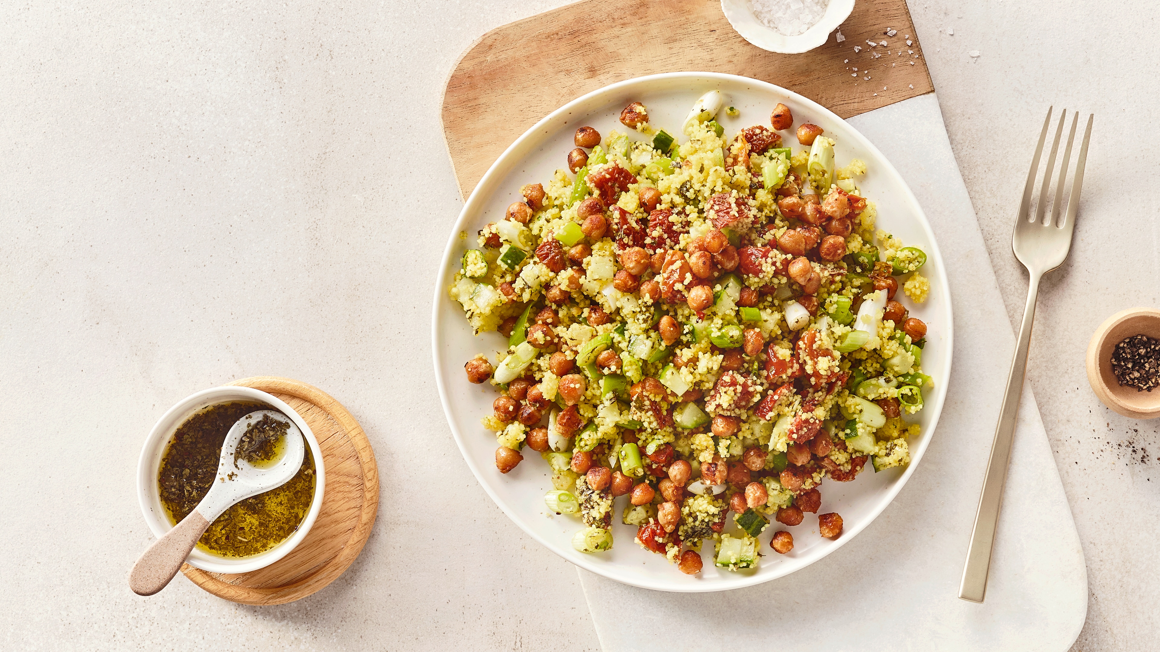 Ein Teller mit Couscous, grünen Gurkenstücken, roten Tomatenstücken und braunen Kichererbsen, daneben eine Schale mit Öl.