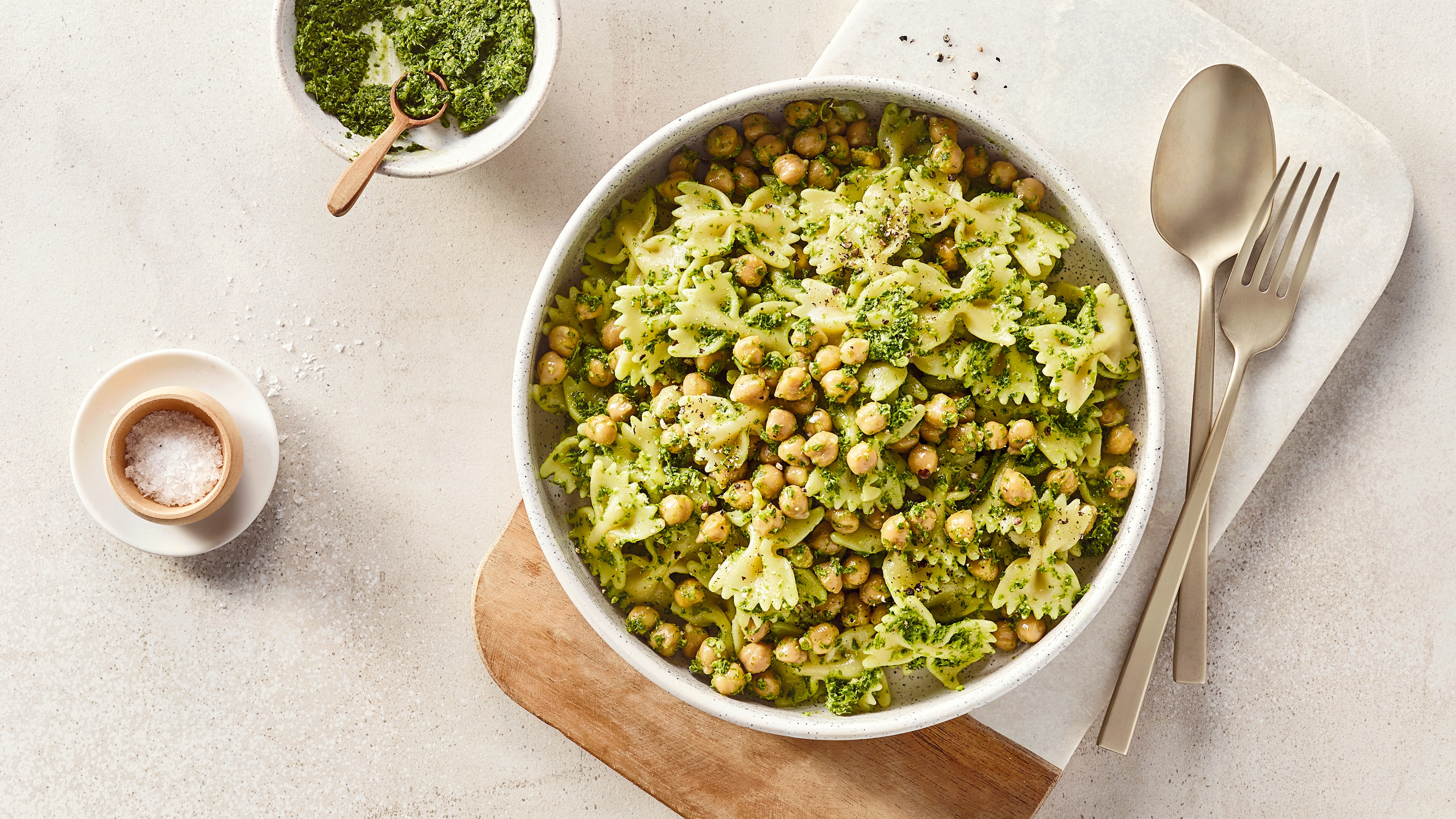 Eine Schüssel mit grüner Farfalle-Nudeln, hellen Kichererbsen & grüner Kräutermischung, daneben eine Schale mit Pesto.