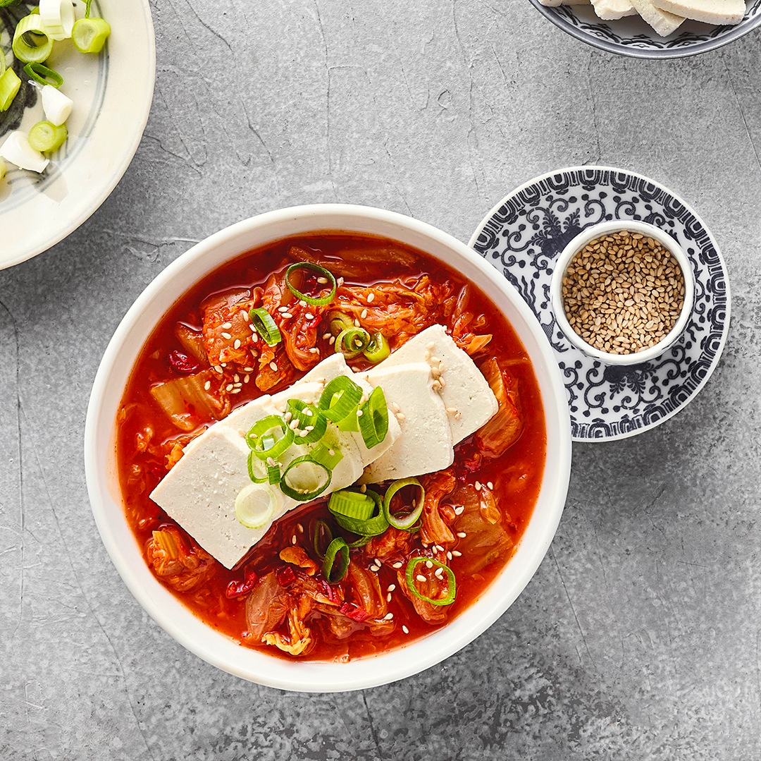 Eine Schale mit roter Suppe, darin Tofustücke und grüne Zwiebeln, daneben kleine Schalen mit Tofu, Sesam und grünen Zwiebeln