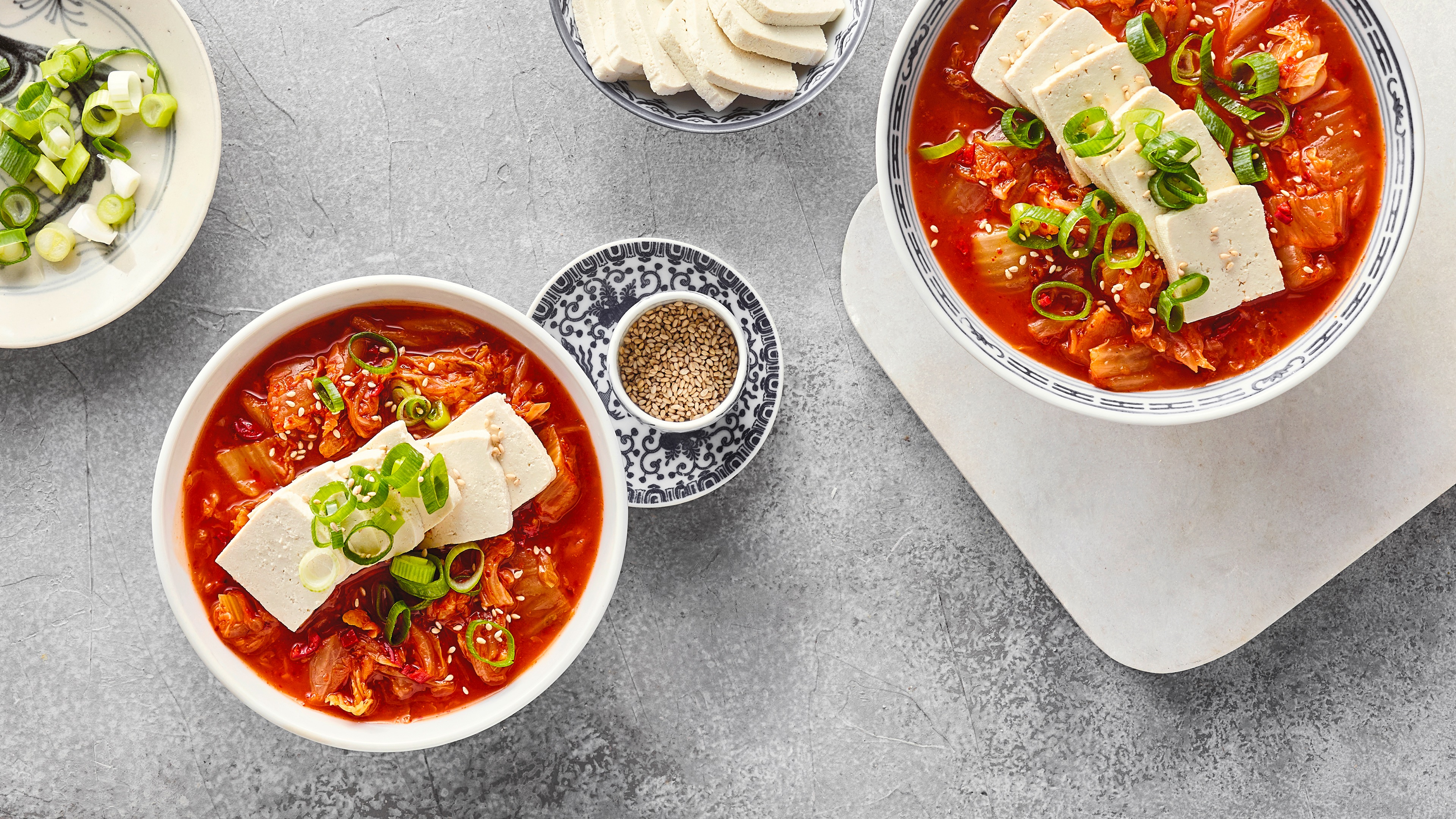 Rezept für Kimchi Jjigae Eine Schale mit roter Suppe, darin Tofustücke und grüne Zwiebeln, daneben kleine Schalen mit Tofu, Sesam und grünen Zwiebeln