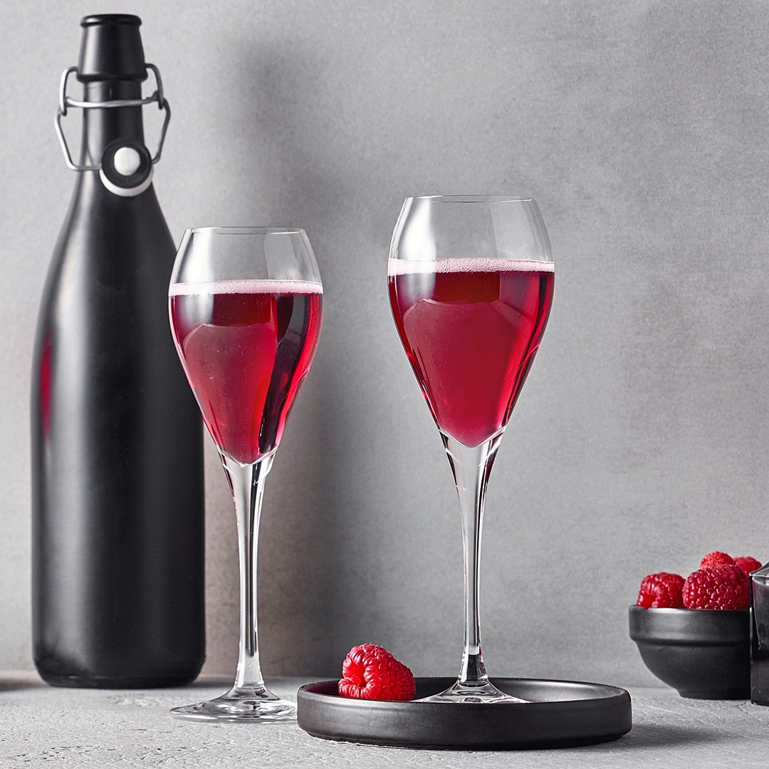 Zwei Gläser mit violettem Getränk, einer schwarzen Flasche dahinter und roten Beeren in kleinen Schalen daneben.