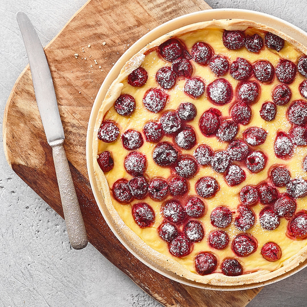 Ein runder Kuchen mit gelber Creme, belegt mit roten Kirschen und bestreut mit weißem Puderzucker.