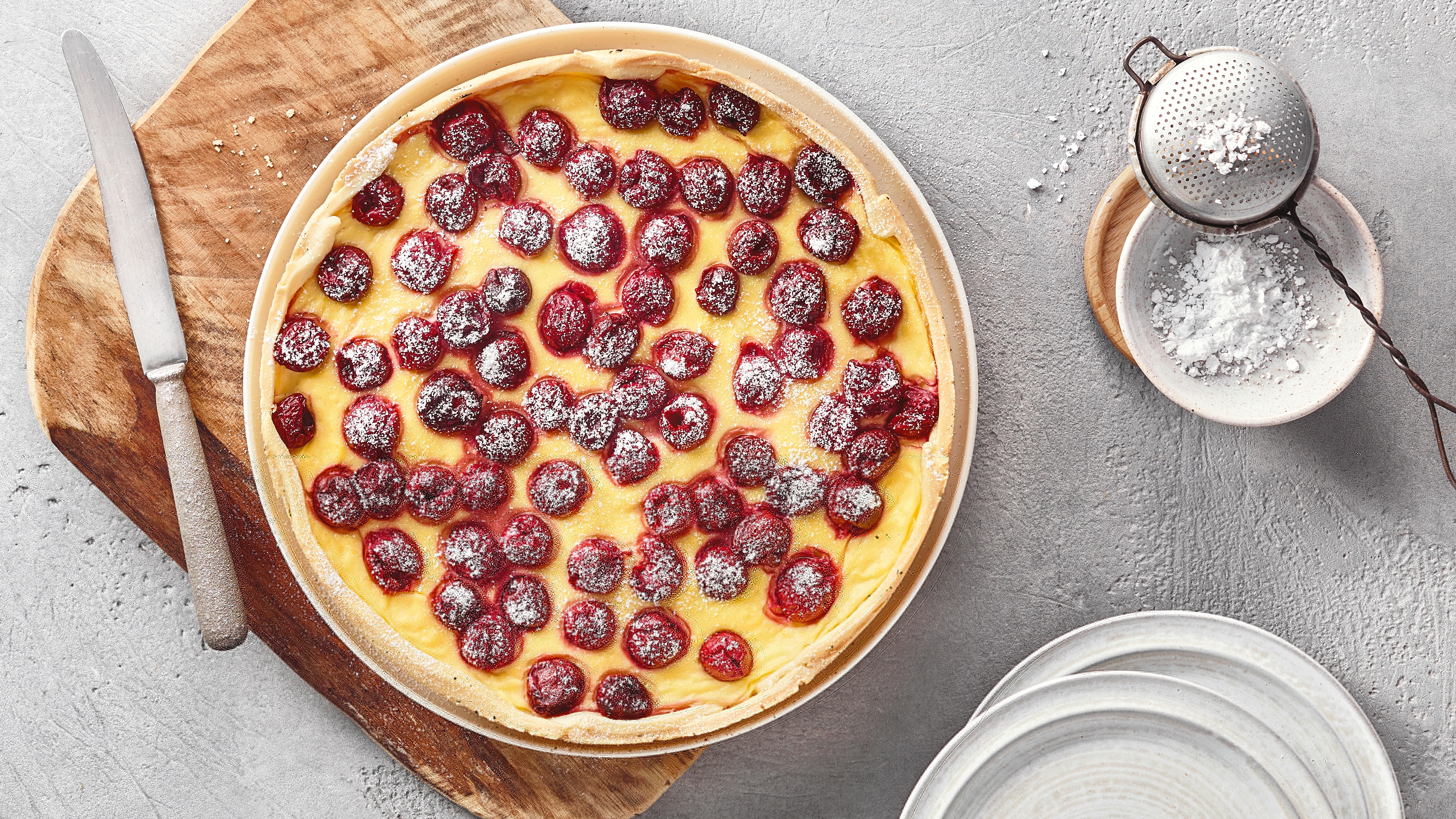Ein runder Kuchen mit gelber Creme, belegt mit roten Kirschen und bestreut mit weißem Puderzucker.