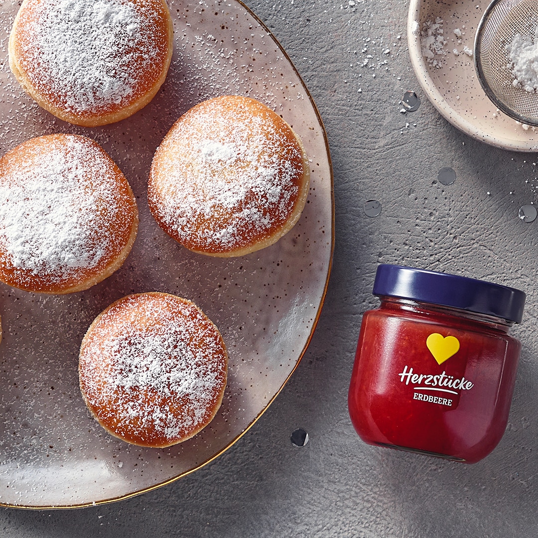 Ein Teller mit sechs runden Berlinern, mit Puderzucker bestreut, daneben eine Schale mit roter Marmelade.