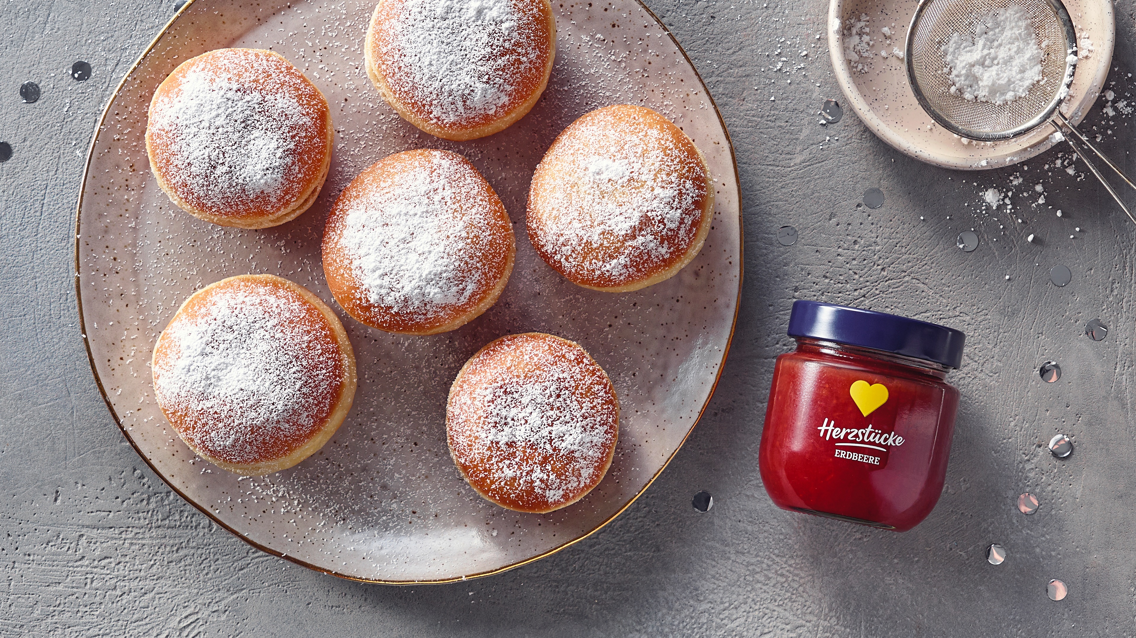 Ein Teller mit sechs runden Berlinern, mit Puderzucker bestreut, daneben eine Schale mit roter Marmelade.