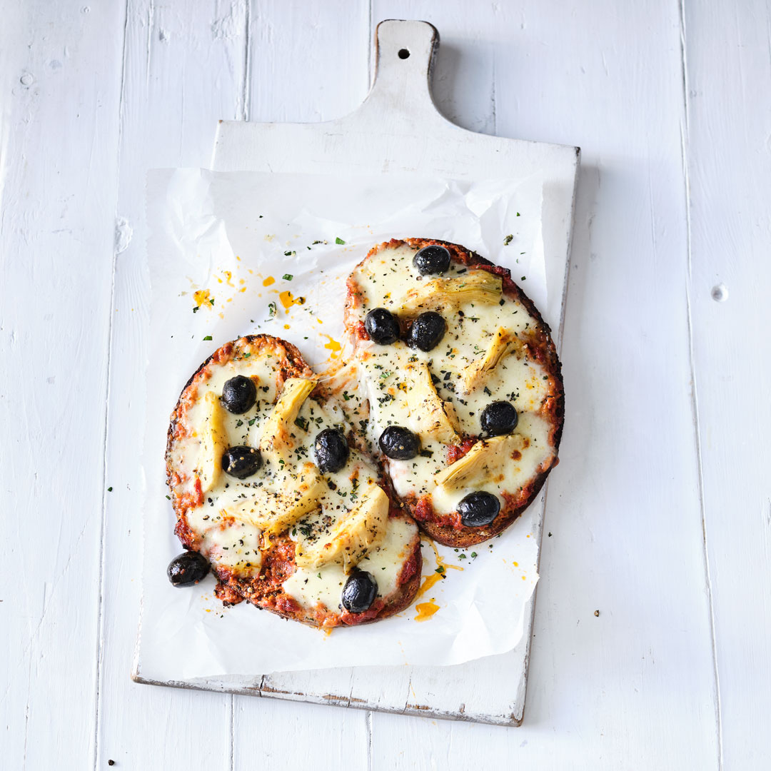 Zwei kleine Pizzen mit geschmolzenem Käse, schwarzen Oliven und Artischockenherzen auf Backpapier.