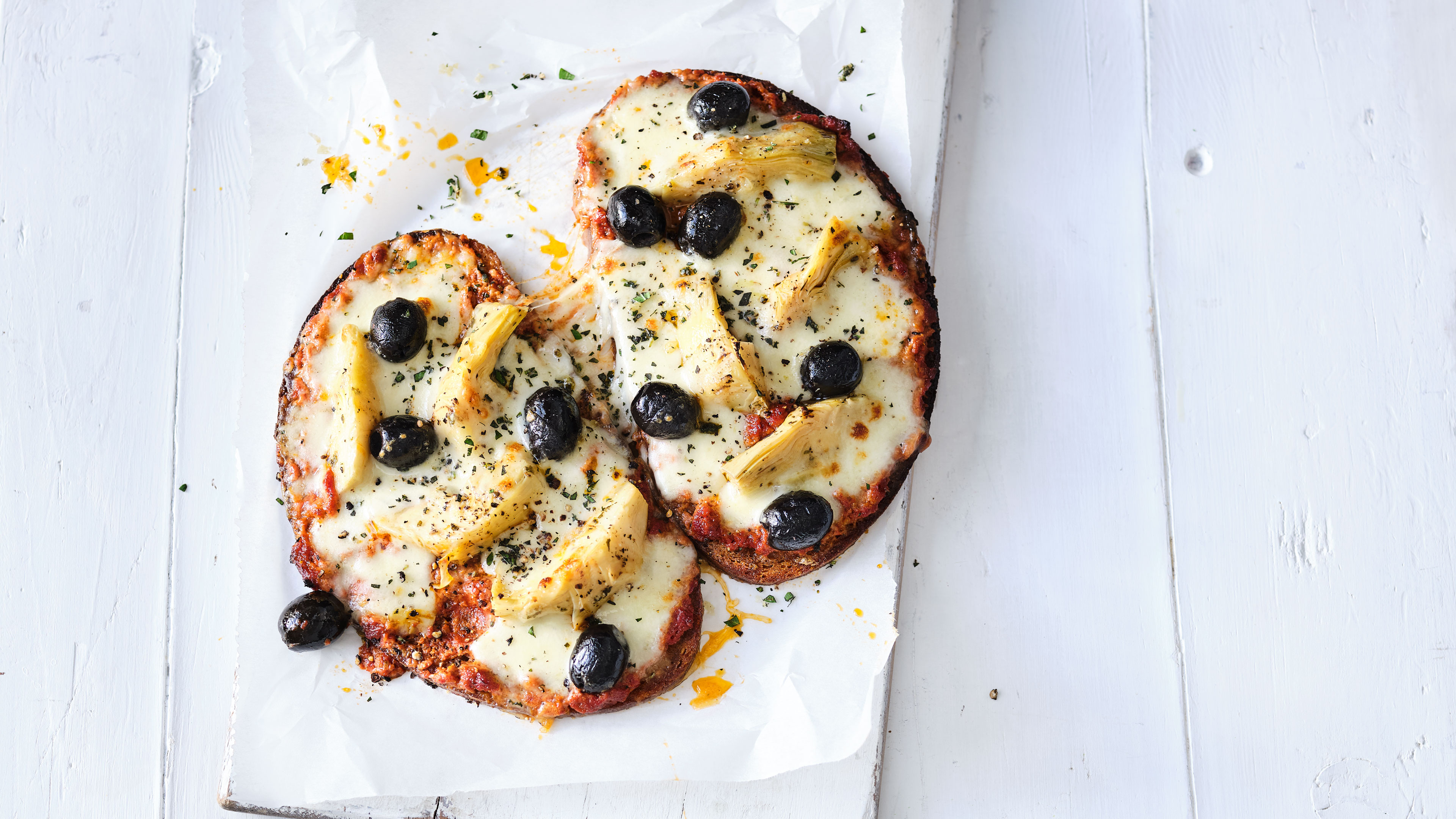 Zwei kleine Pizzen mit geschmolzenem Käse, schwarzen Oliven und Artischockenherzen auf Backpapier.