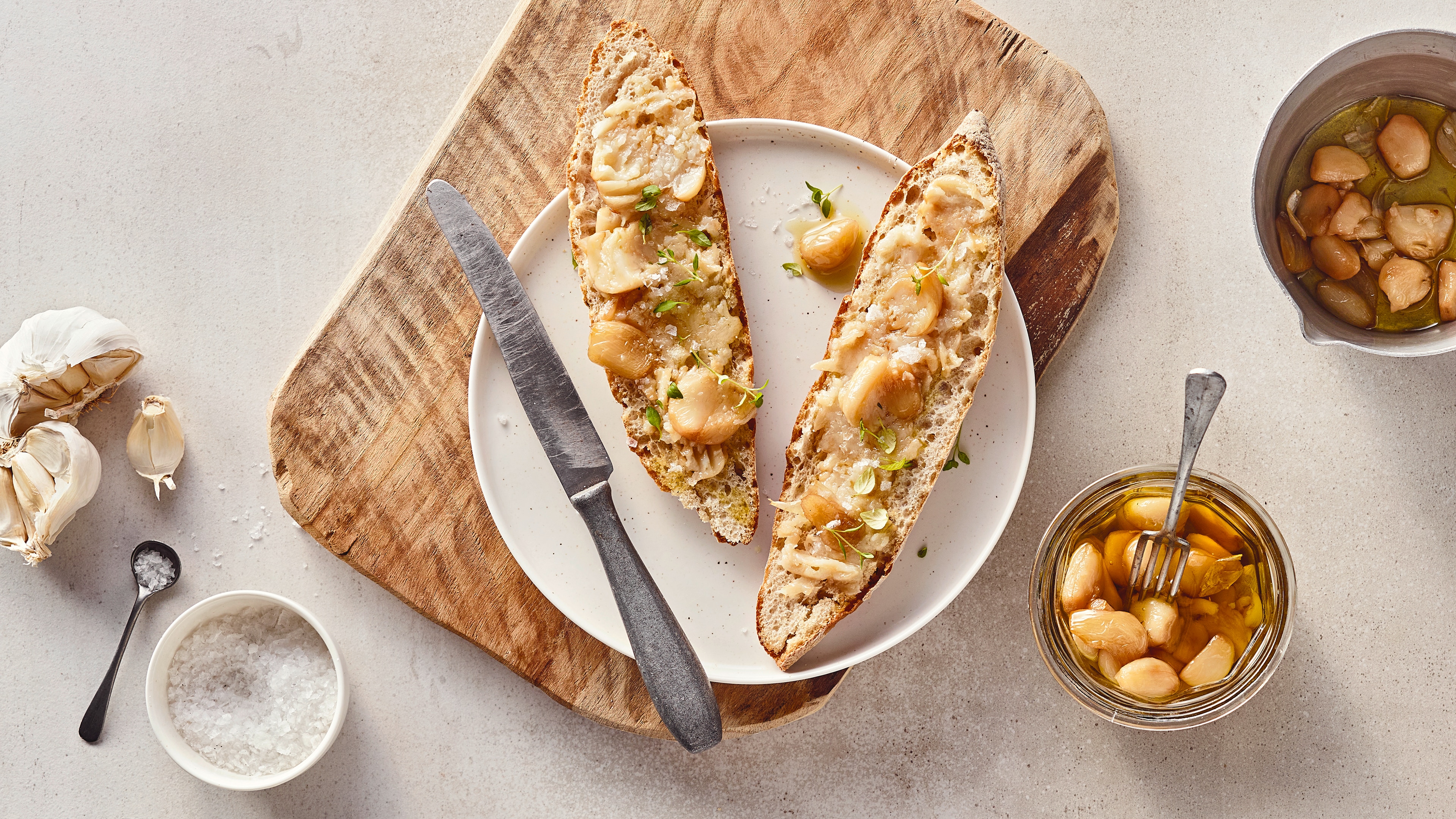 Rezept für Knoblauch-Confit Ein Teller mit länglichen Brötchenhälften, bestrichen mit zerdrücktem Knoblauch, daneben ein Gläschen mit Knoblauch in Öl