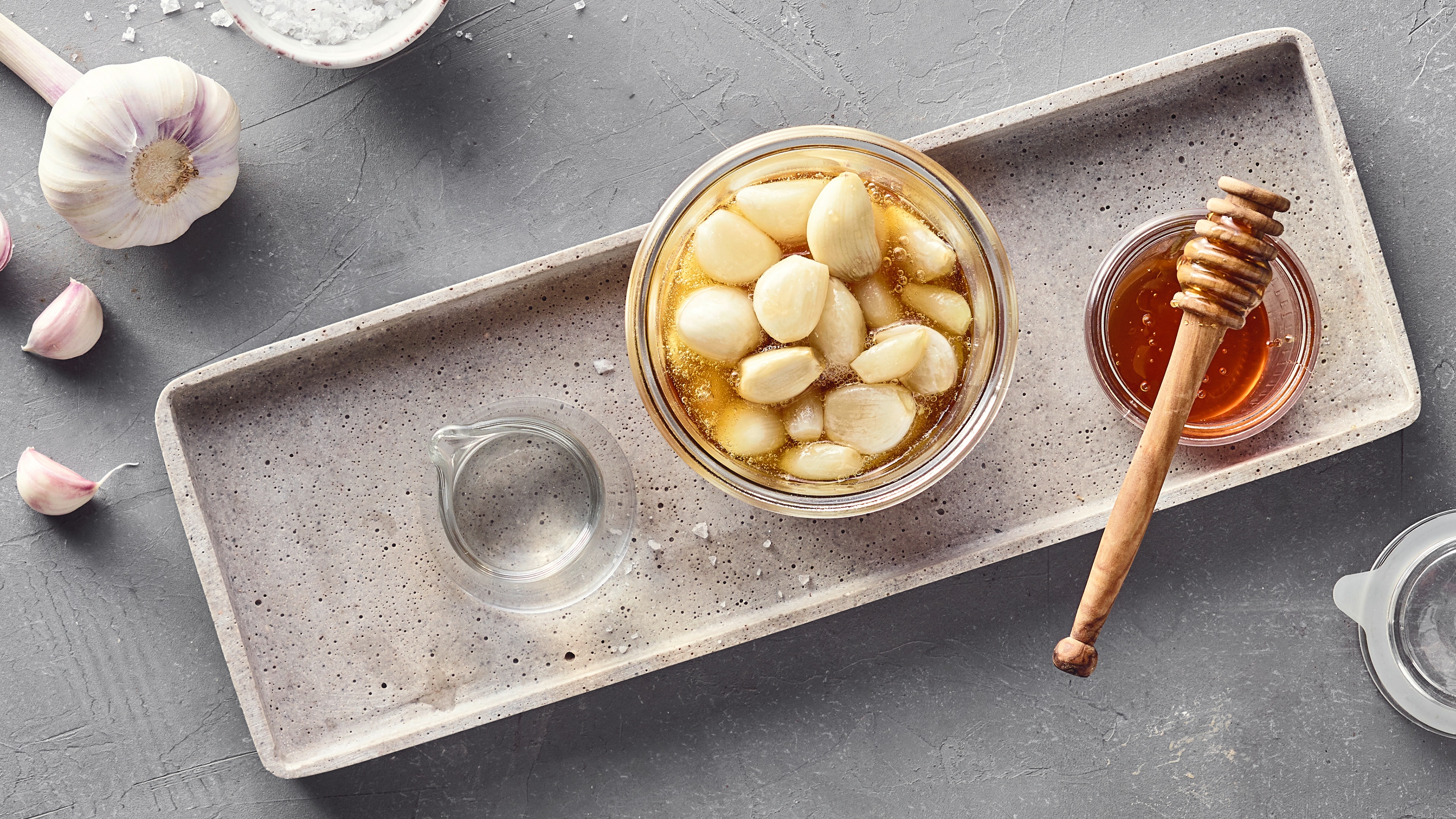 Rezept für fermentierten Knoblauch Ein Glas mit geschälten Knoblauchzehen in heller Flüssigkeit, daneben eine Schale mit Honig und grobem Salz