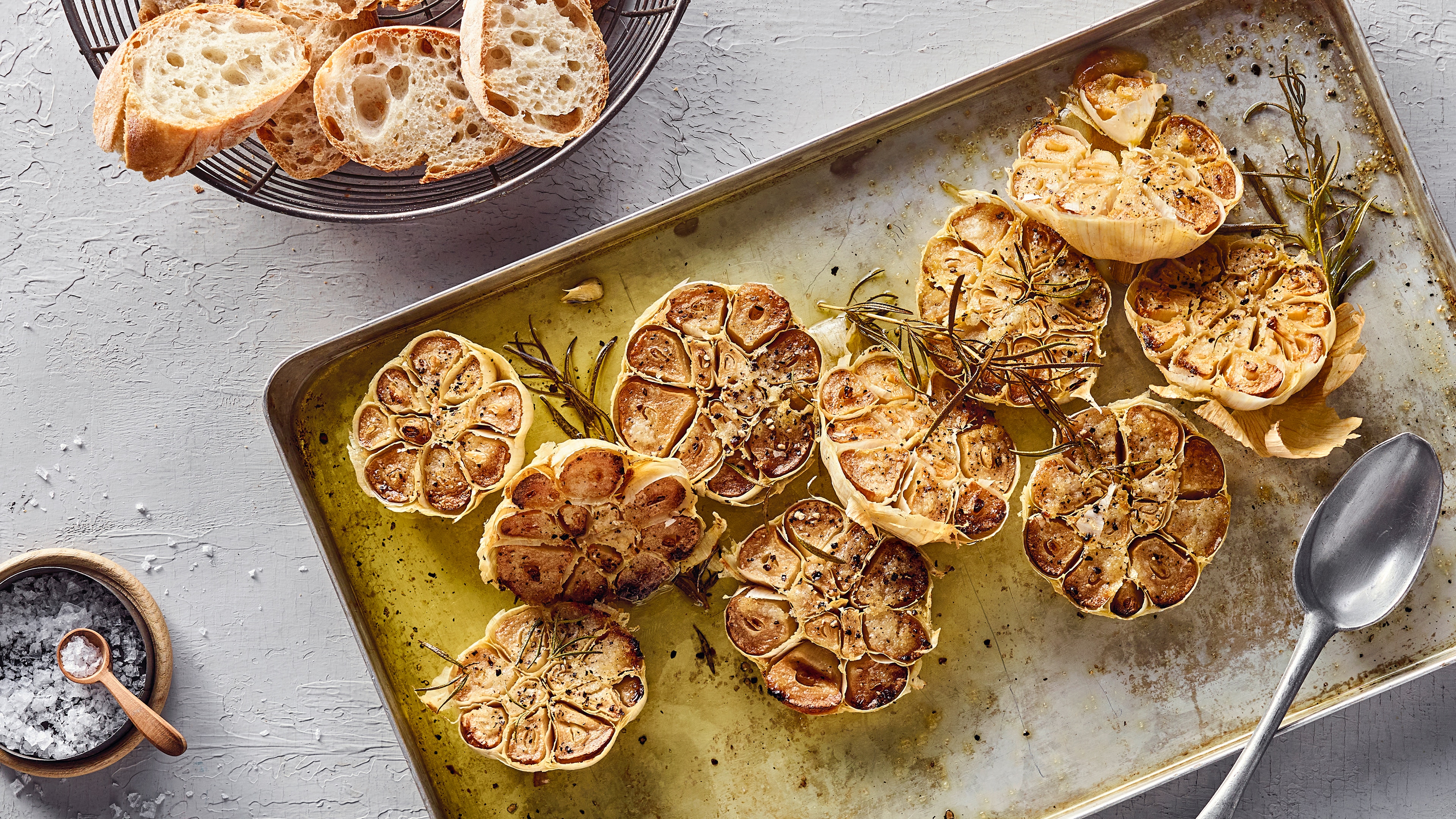 Rezept für Knoblauch im Ofen Ein eisernes Backblech mit braun gebackenen, saftigen Knoblauchhälften, getoppt mit Öl und Rosmarin, daneben ein Korb mit frischem Brot