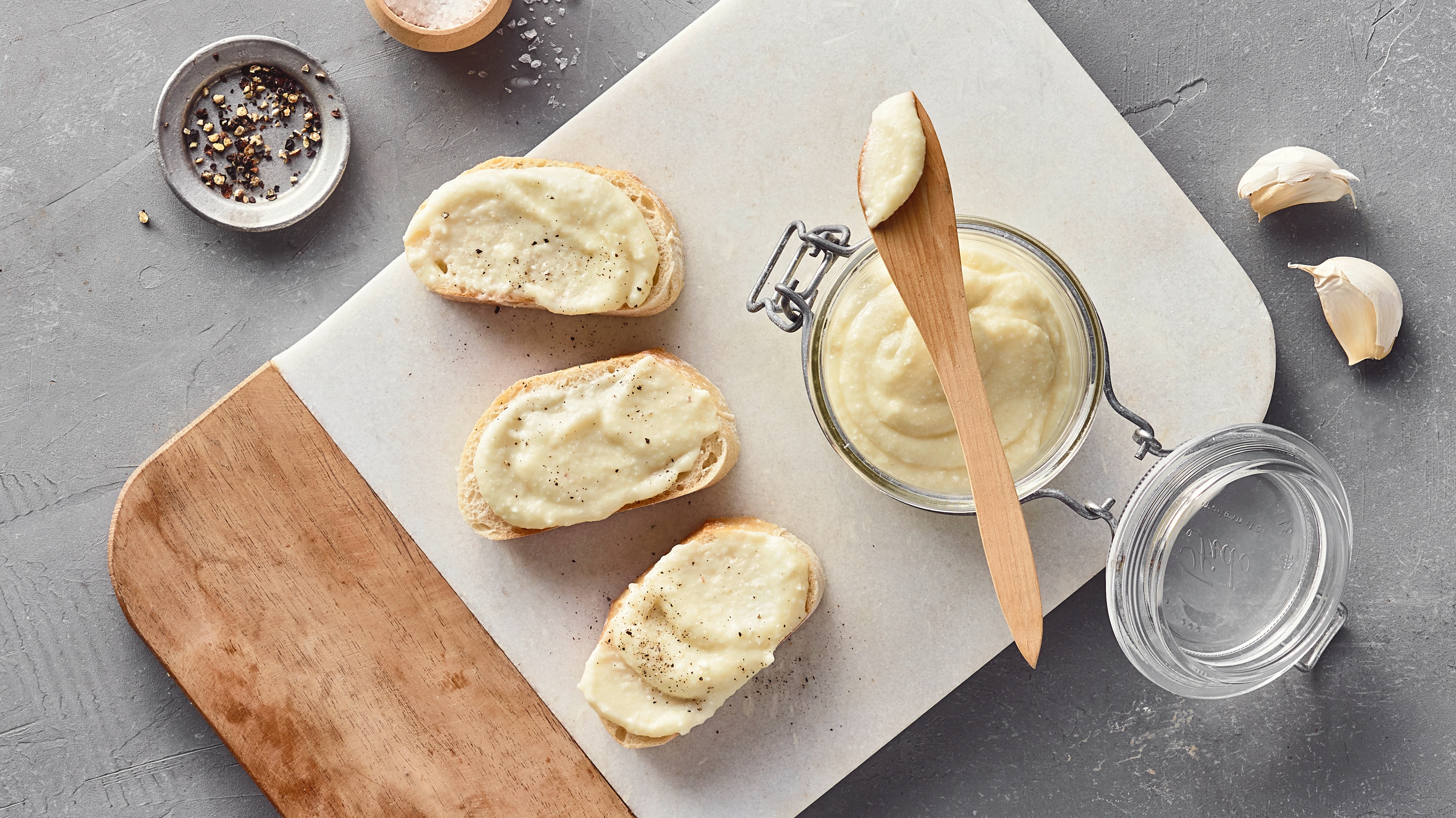 Rezept für Knoblauchpaste Ein Glas mit weißem cremigem Aufstrich, daneben drei Brotscheiben mit dem gleichen Aufstrich und ein Holzlöffel