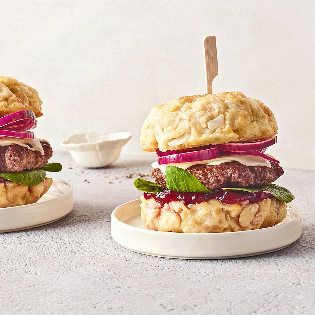 Zwei Burger mit Rindfleisch-Patty, roten Zwiebelringen, Salat und heller Sauce zwischen Knödel-Brötchen auf kleinen Tellern.
