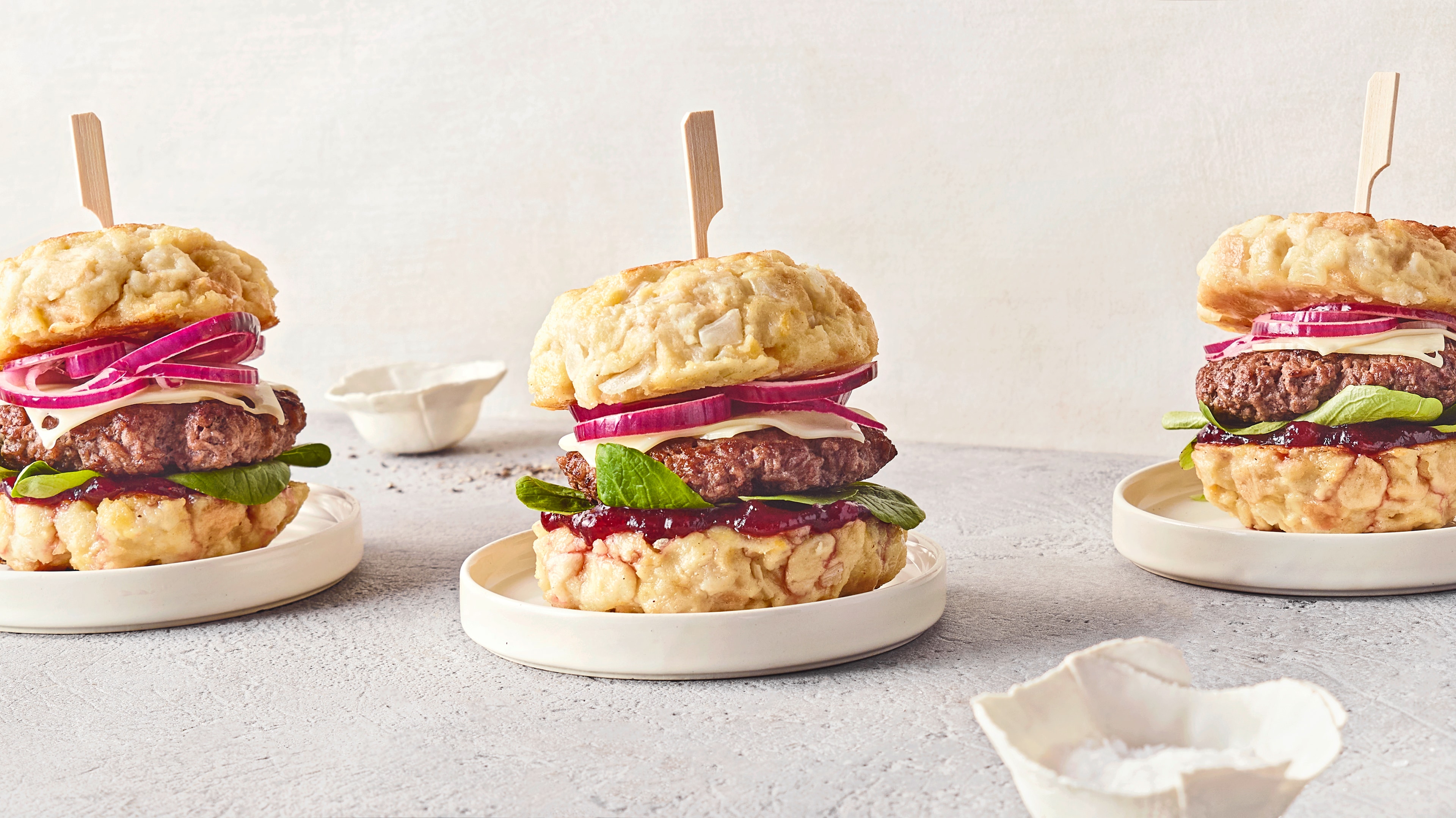 Drei Burger mit Rindfleisch-Patty, roten Zwiebelringen, Salat und heller Sauce zwischen Knödel-Brötchen auf kleinen Tellern.