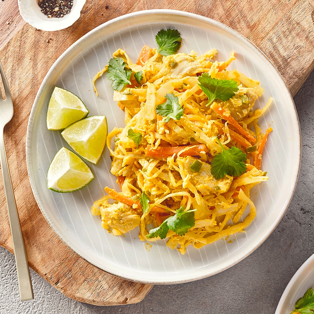 Ein Teller mit gelben Nudeln, orangefarbenen Streifen, hellen Fleischstücken, grünen Kräutern und drei Stücken grüner Limette.
