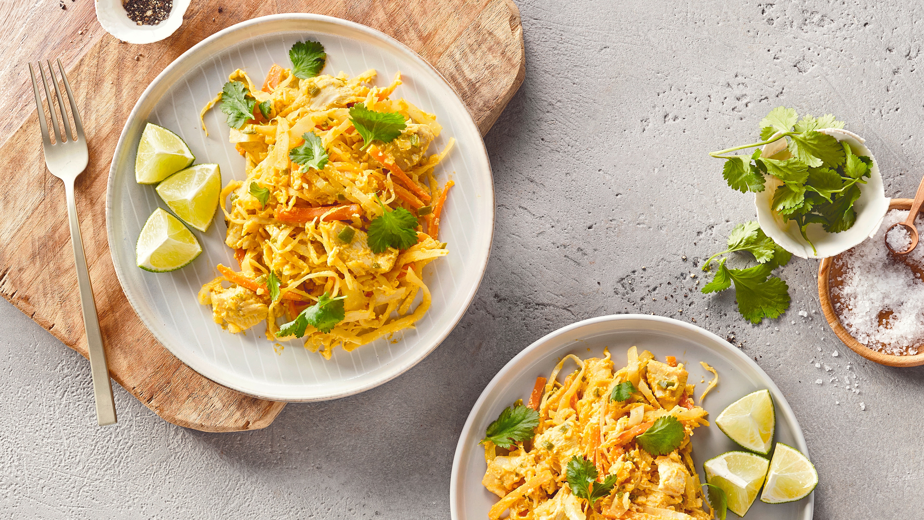 Ein Teller mit gelben Nudeln, orangefarbenen Streifen, hellen Fleischstücken, grünen Kräutern und drei Stücken grüner Limette.