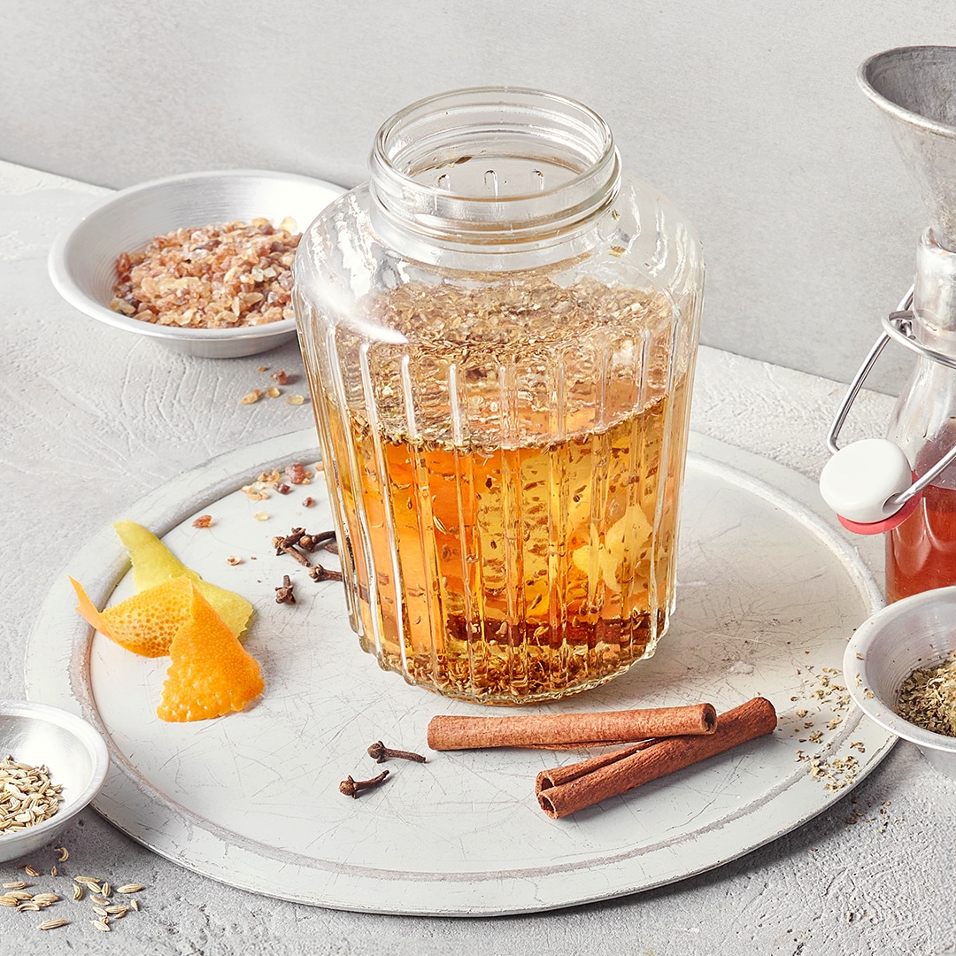 Ein Glas mit goldener Flüssigkeit, darin braune Zimtstangen, helle Zitruszesten, dunkle Nelken und feine Kräuter.
