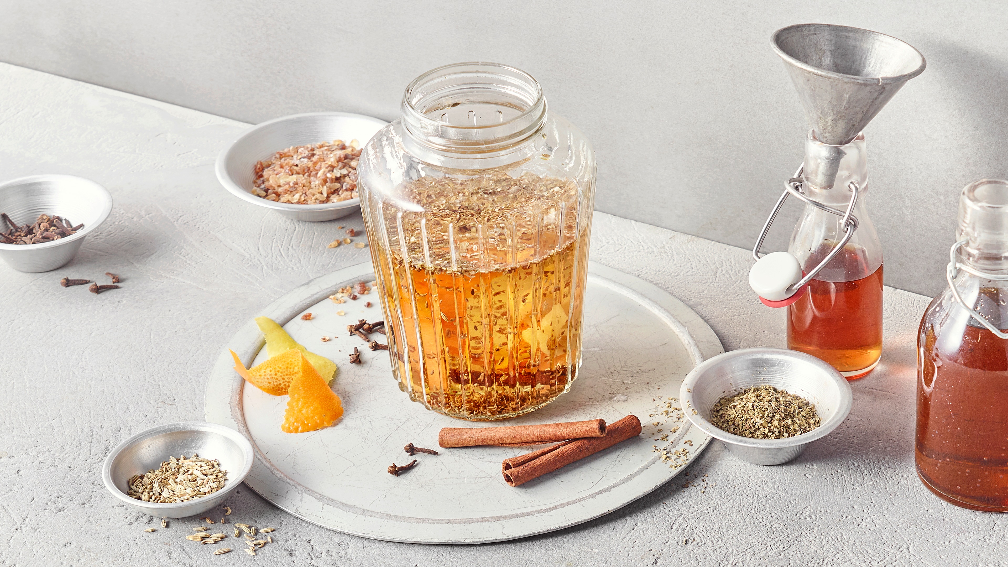 Ein Glas mit goldener Flüssigkeit, darin braune Zimtstangen, helle Zitruszesten, dunkle Nelken und feine Kräuter.