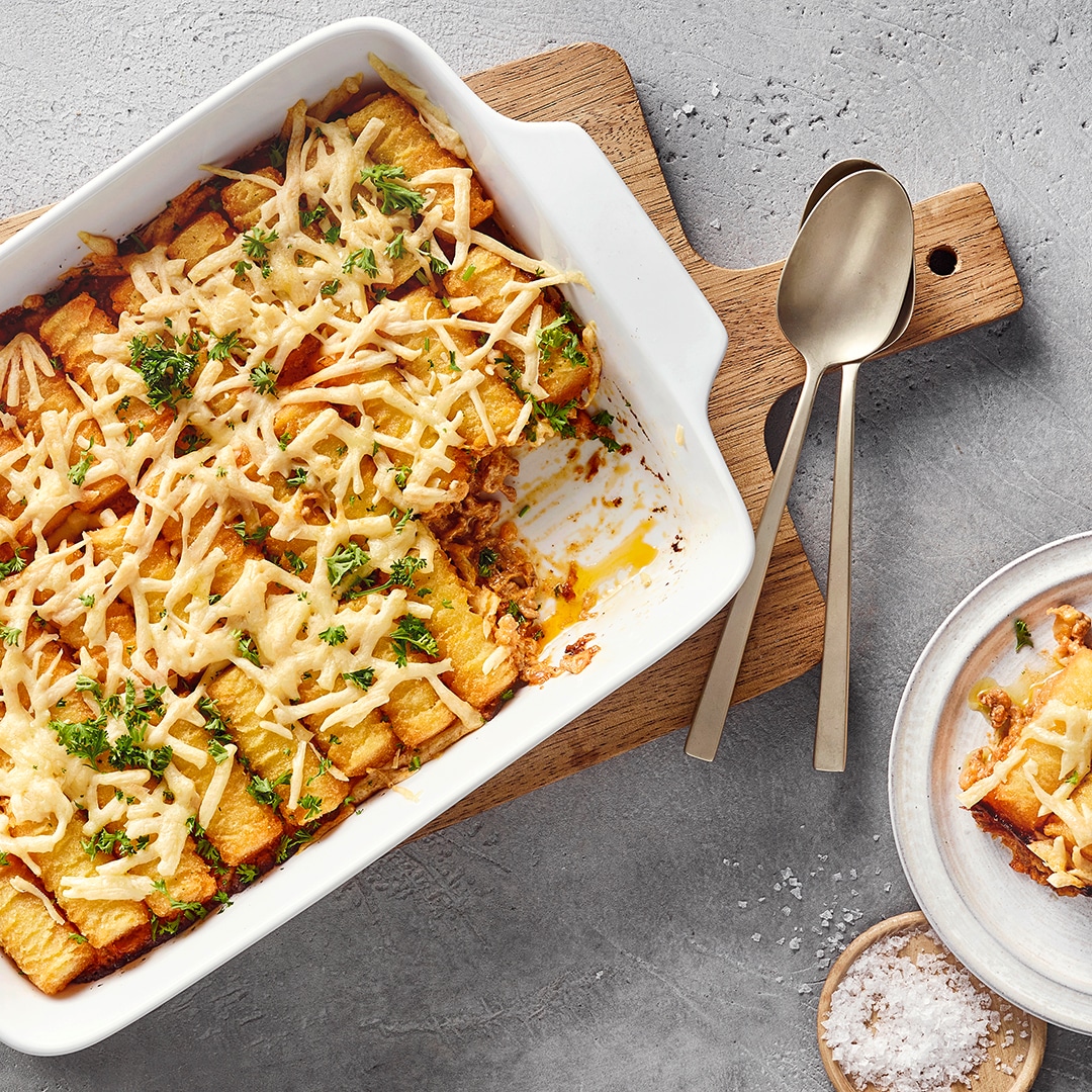 Eine Auflaufform mit Kroketten, überbackenem weißem Käse und grünen Kräutern, daneben ein Teller mit einer Portion.