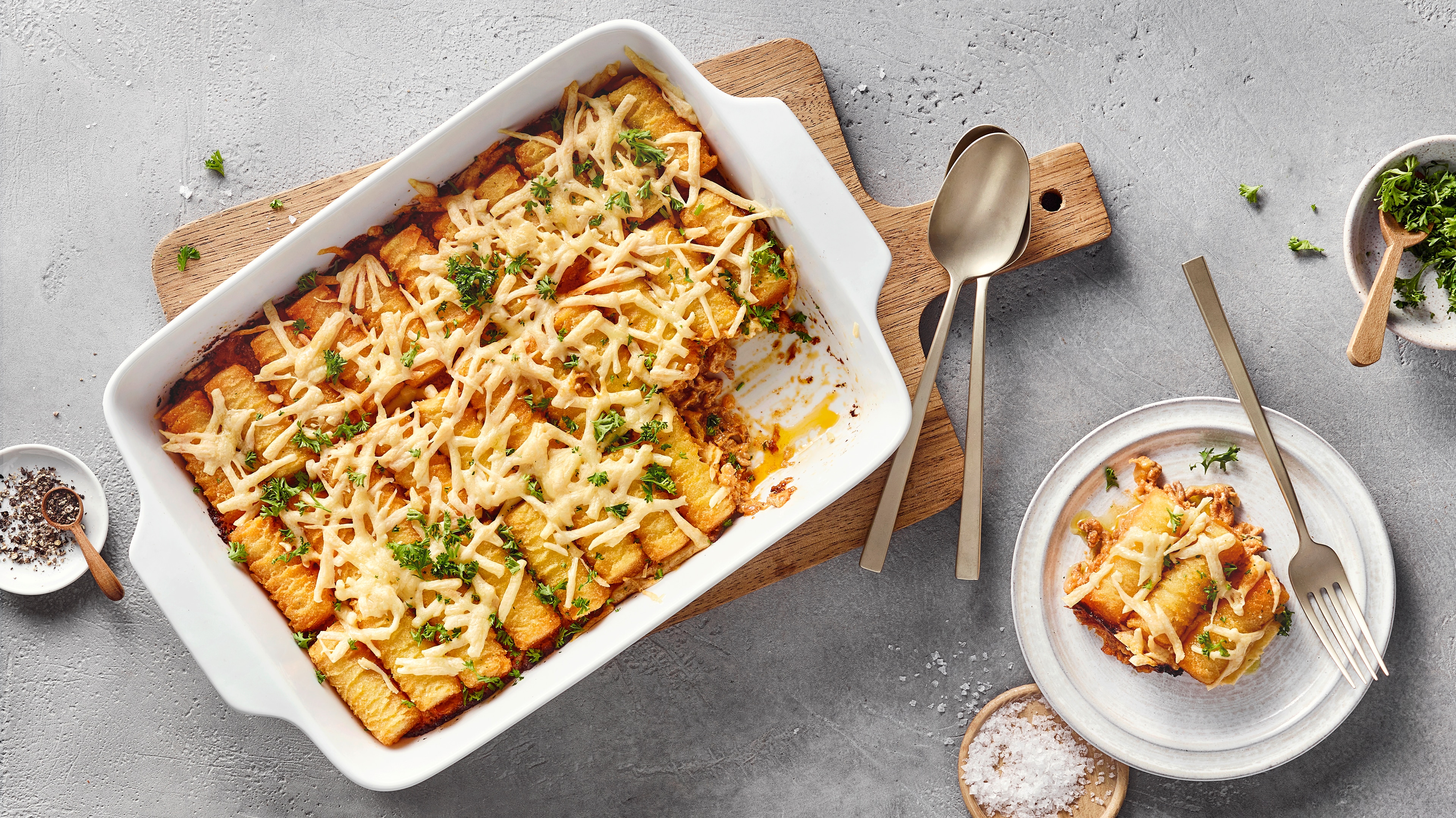 Eine Auflaufform mit Kroketten, überbackenem weißem Käse und grünen Kräutern, daneben ein Teller mit einer Portion.