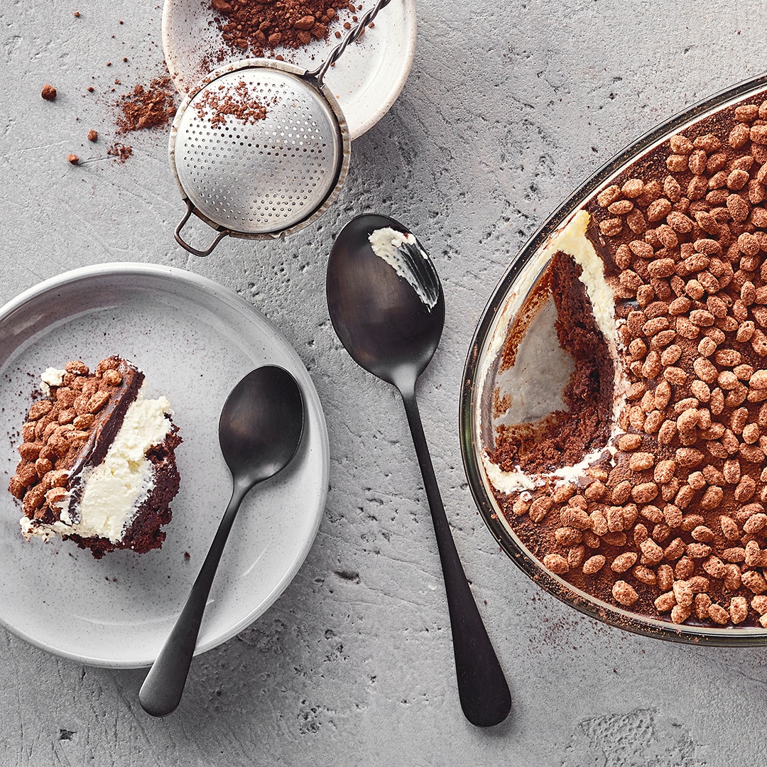 Ein Teller mit einem Stück Schichtkuchen aus Schokolade und Creme, bestreut mit Kakaopulver und knusprigen Schokoflocken