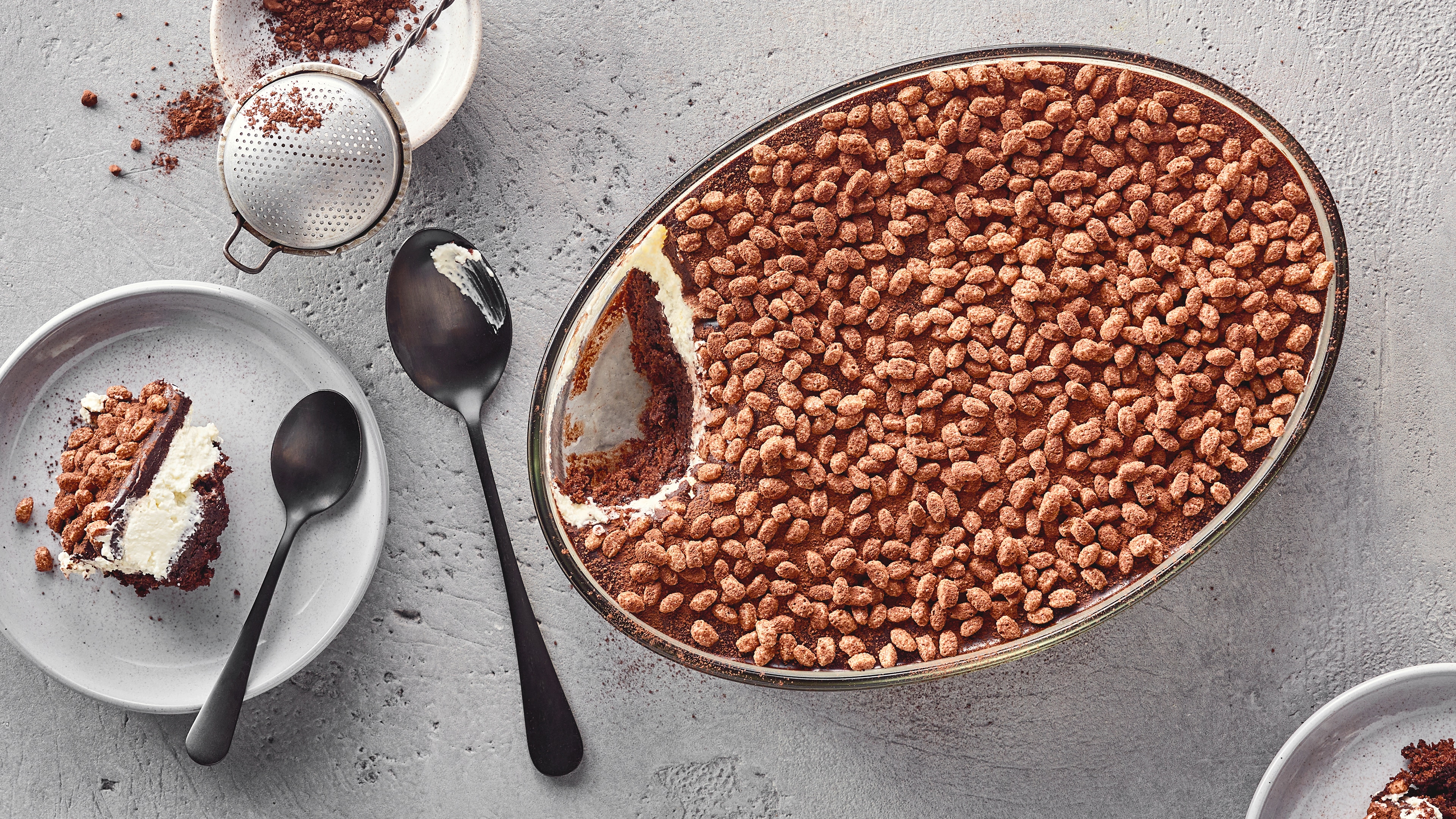 Rezept für Krunchoco Cake Ein Teller mit einem Stück Schichtkuchen aus Schokolade und Creme, bestreut mit Kakaopulver und knusprigen Schokoflocken