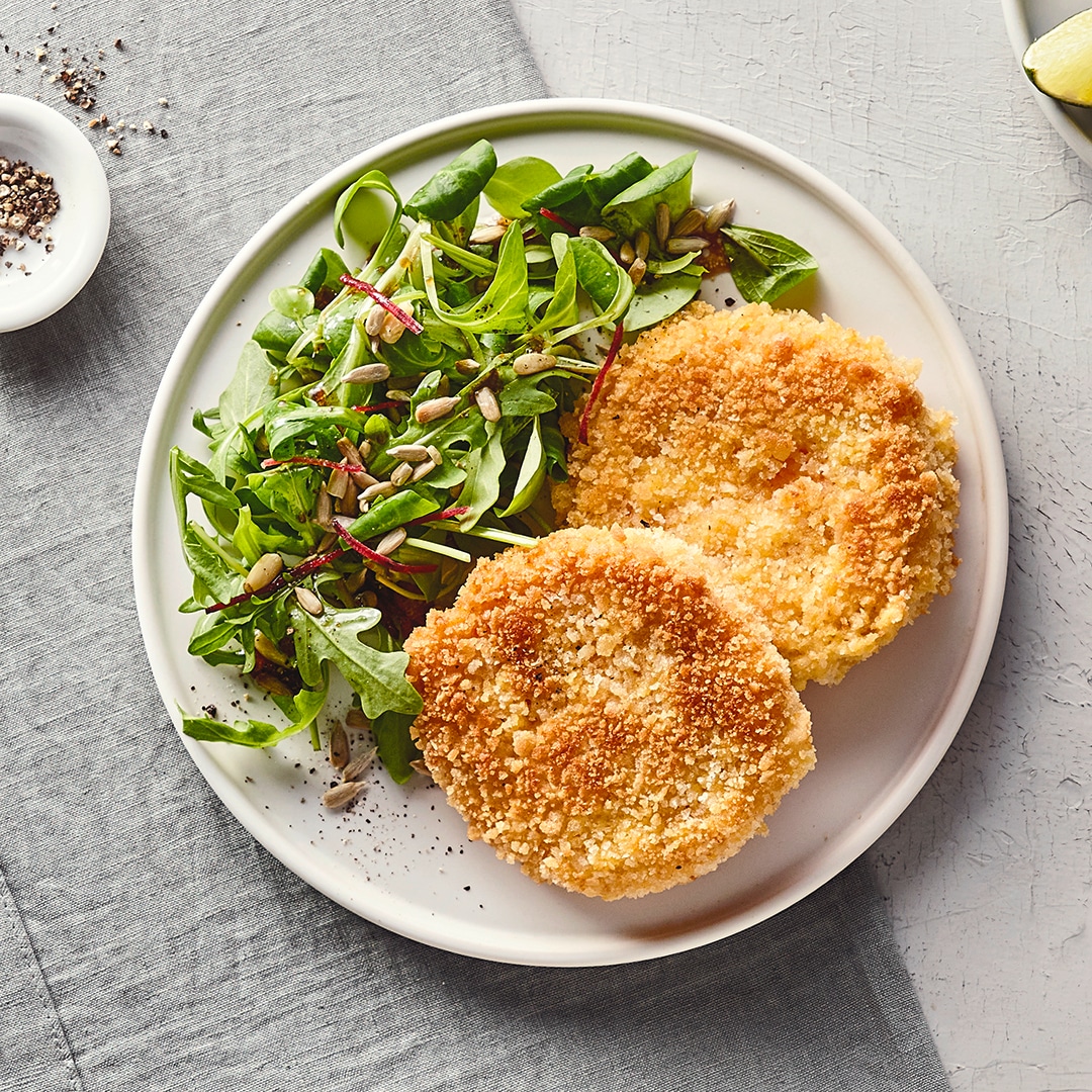 Ein großer Teller mit zwei panierten, knusprigen Talern und einem Salat aus Feldsalat und Rucola, getoppt mit Körnern