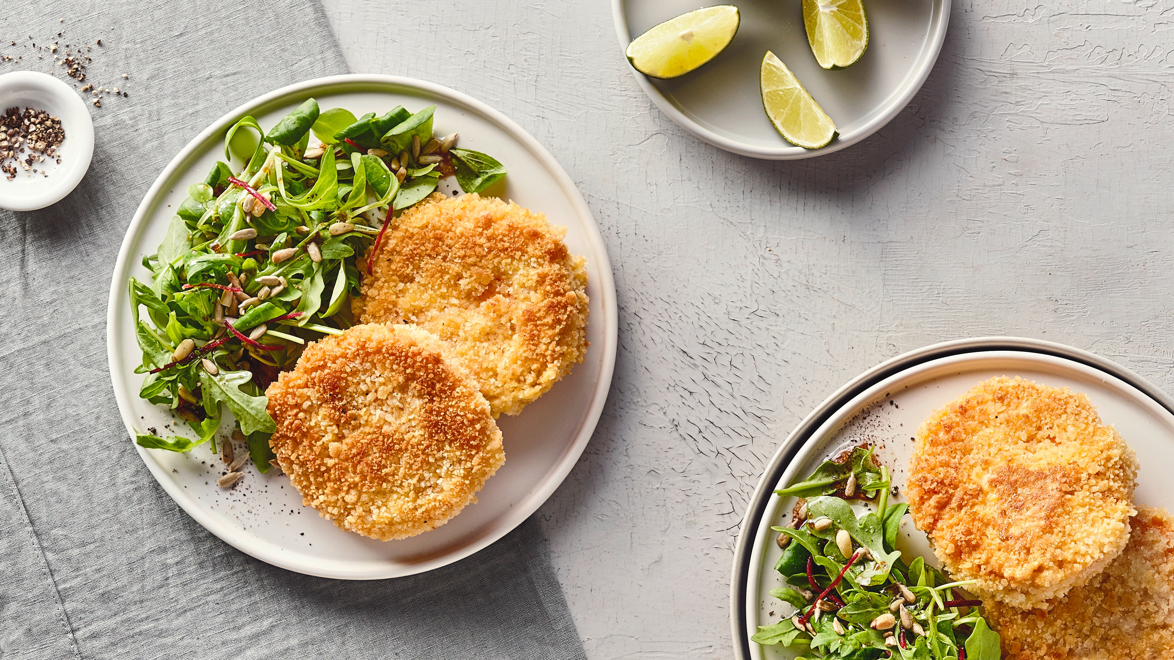 Rezept für Kürbisschnitzel Ein großer Teller mit zwei panierten, knusprigen Talern und einem Salat aus Feldsalat und Rucola, getoppt mit Körnern