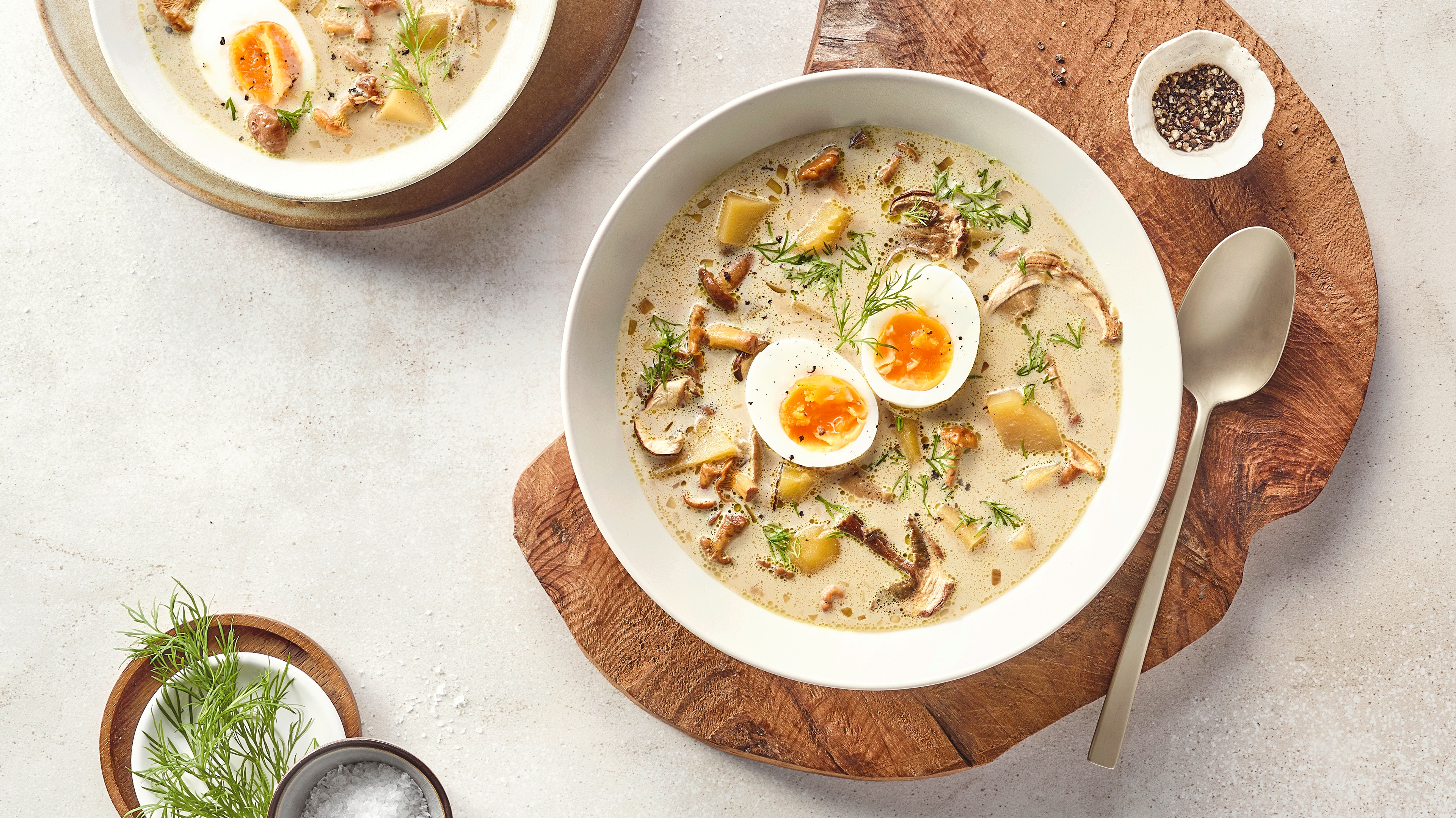Ein Teller mit heller cremiger Suppe, darin Kartoffelstücke, Pilzscheiben, Dill und einem halbierten weich gekochten Ei.