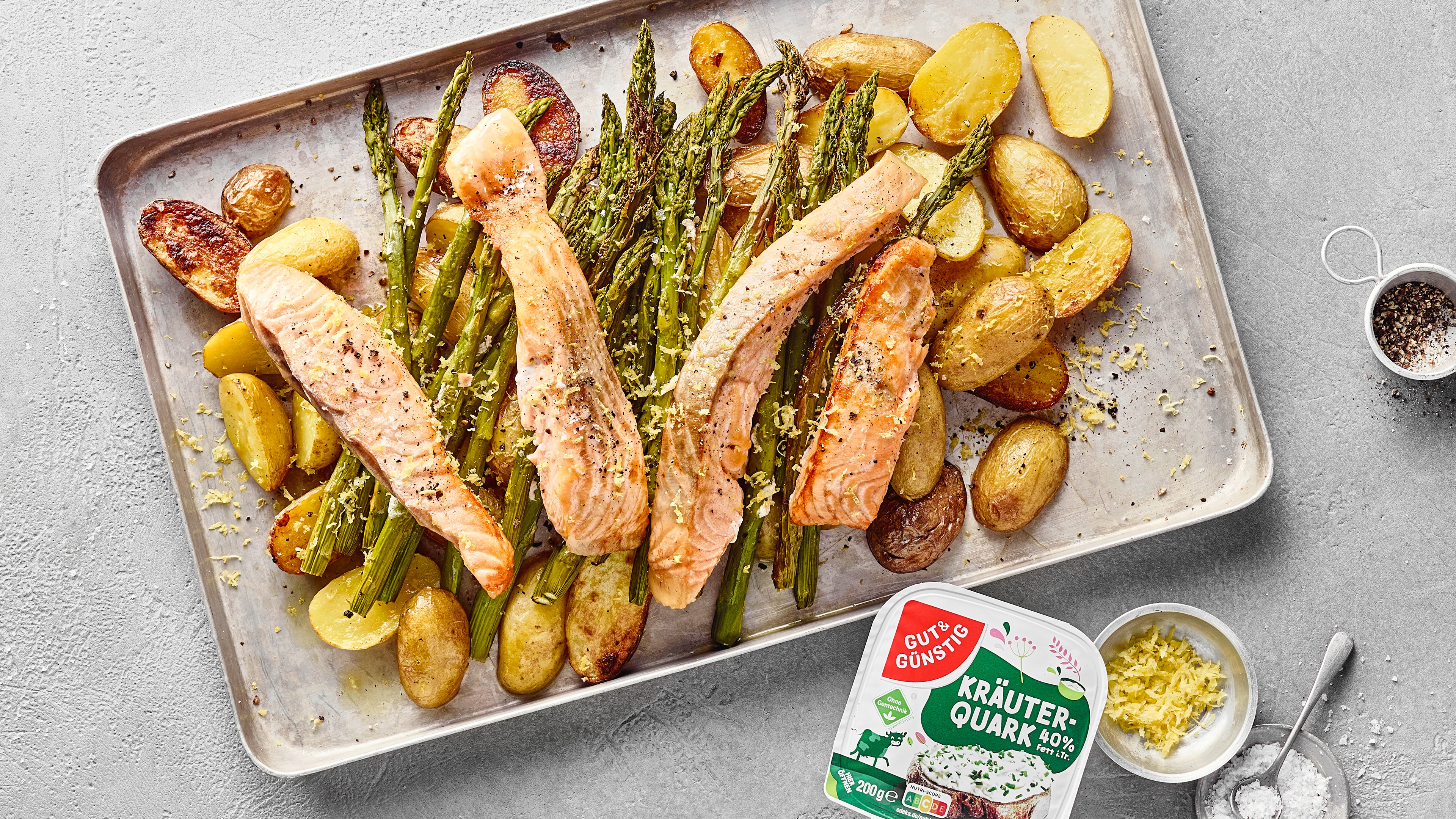 Ofenblech mit Lachsfilets, umgeben von goldbraunen Kartoffelhälften und grünem Spargel. Daneben GUT&GüNSTIG Kräuterquark.