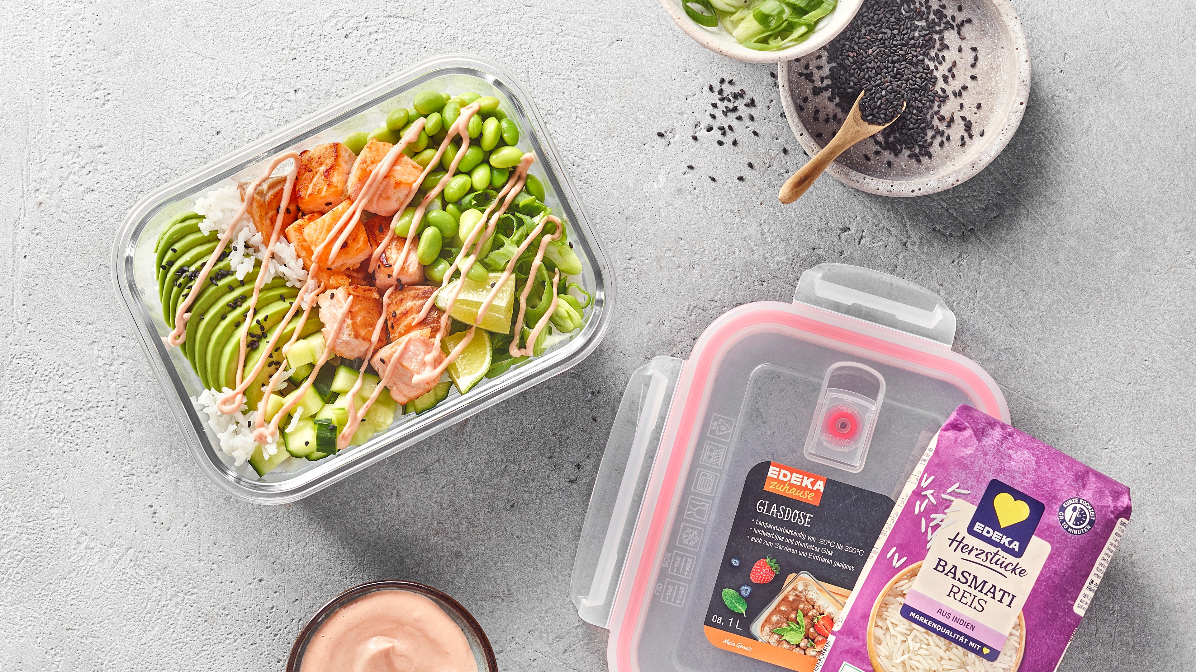 Meal-Prep-Box mit Basmatireis, Avocado, Edamame und gebratenen Hähnchenstücken, daneben eine Packung EDEKA Herzstücke Basmatireis und EDEKA Zuhause Glasdose.