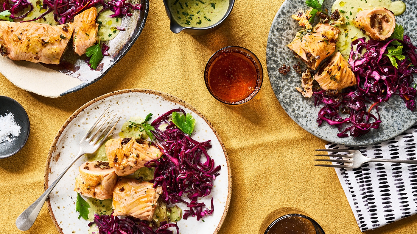 Mit Pumpernickel gefüllte Lachsröllchen mit Rotkohlsalat und cremiger Soße auf mehreren Tellern.