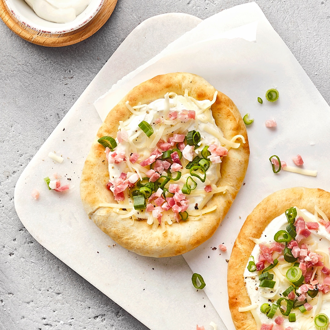 Brett mit zwei Fladenbroten auf einem Papiertuch. Die Brote sind mit Sauerrahm, Frühlingszwiebeln und Speckwürfeln belegt.