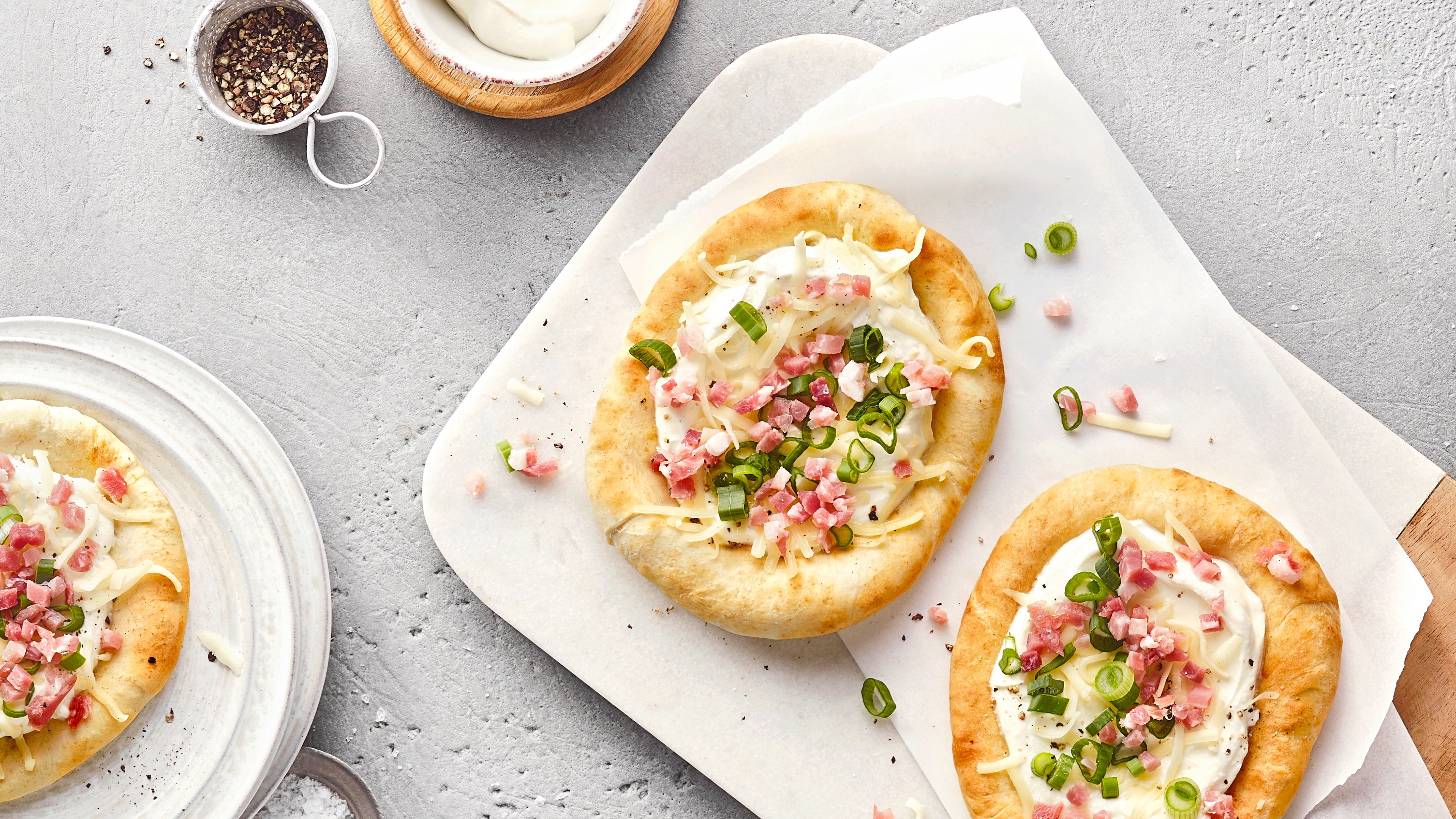 Brett mit zwei Fladenbroten auf einem Papiertuch. Die Brote sind mit Sauerrahm, Frühlingszwiebeln und Speckwürfeln belegt. Links unten ist ein weiteres Fladenbrot teilweise zu sehen.