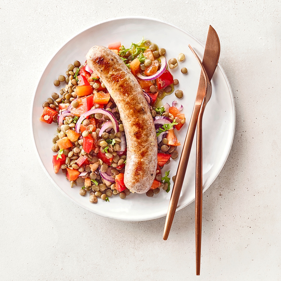Teller mit Linsen-Gemüse-Salat und gebratener Bratwurst, angerichtet mit Tomaten, roten Zwiebeln und Kräutern.
