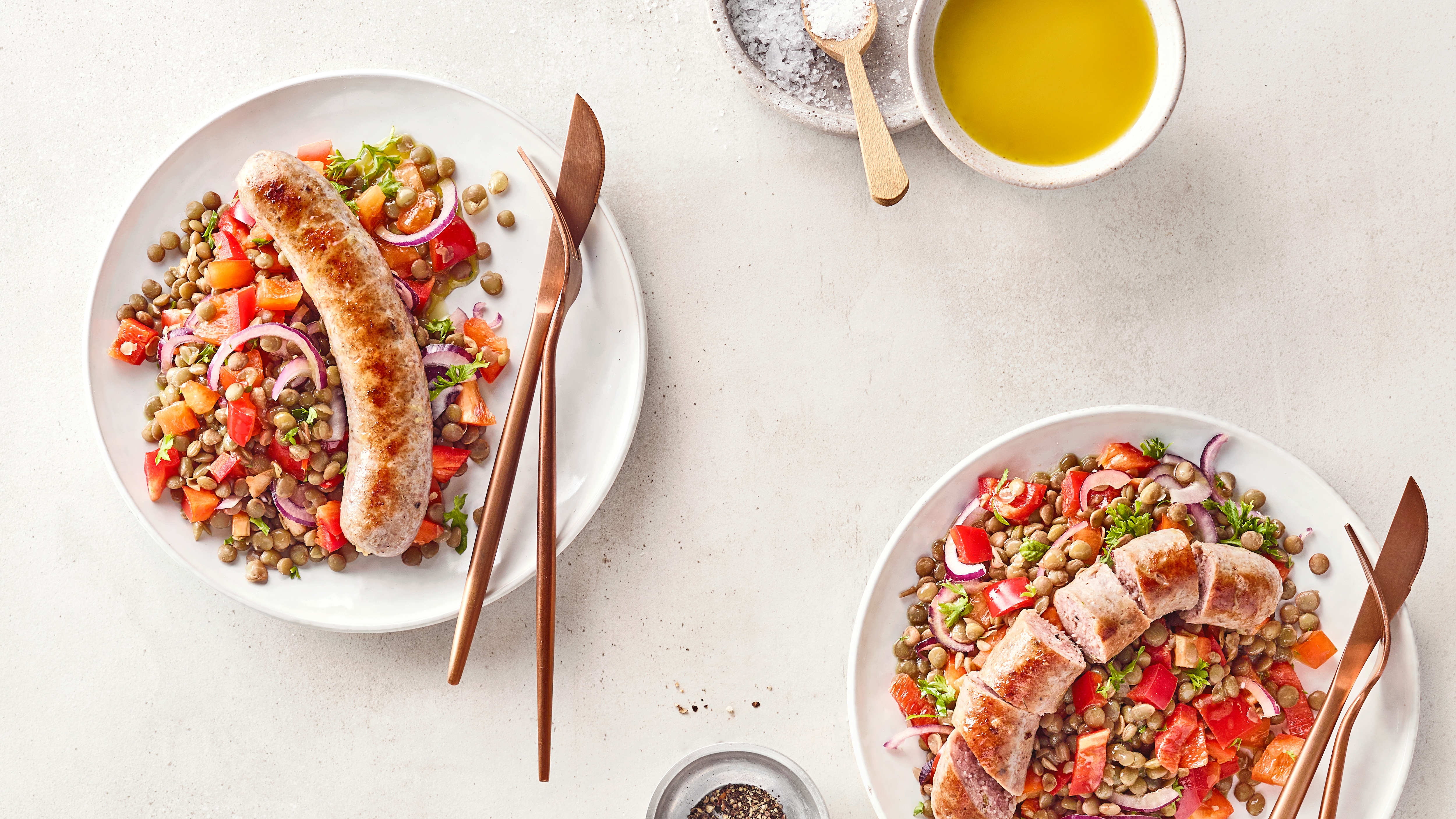 Zwei Teller mit Linsen-Gemüse-Salat und gebratener Bratwurst, angerichtet mit Tomaten, roten Zwiebeln und Kräutern.