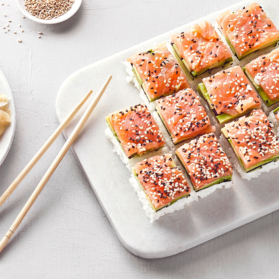 Blech mit mehreren quadratischen Sushi-Stücken aus Lachs, Avocado, Reis und Sesam. Daneben liegen Essstäbchen.